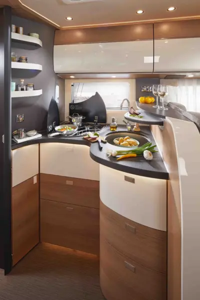 Moderne Carthago C Tourer Wohnmobilküche mit Holz- und weißen Schränken, schwarzen Arbeitsplatten und eingebauten Regalen. Frisches Gemüse, Kräuter und Geschirr stehen auf der Theke, im Hintergrund stehen Weingläser und Obst. Durchgehend warme Raumbeleuchtung.