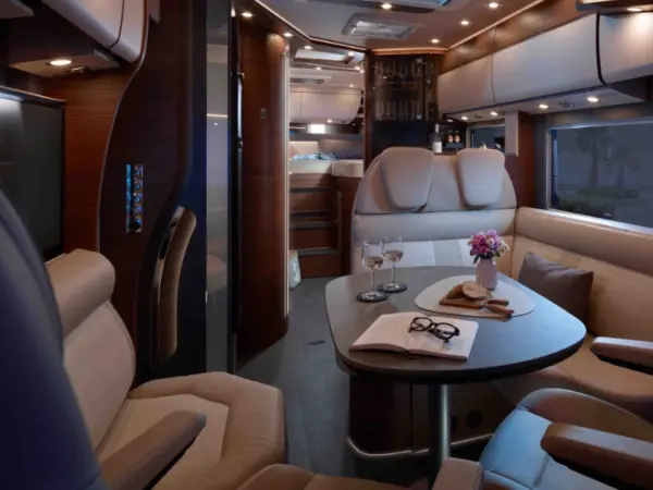 Luxuriöses Carthago Chic C Line T Wohnmobil-Interieur mit beigen Ledersitzen, einem Esstisch mit Weingläsern, einem Buch und Blumen. Die warme Beleuchtung unterstreicht die Holzverkleidung und die modernen Annehmlichkeiten für eine gemütliche, gehobene Atmosphäre.