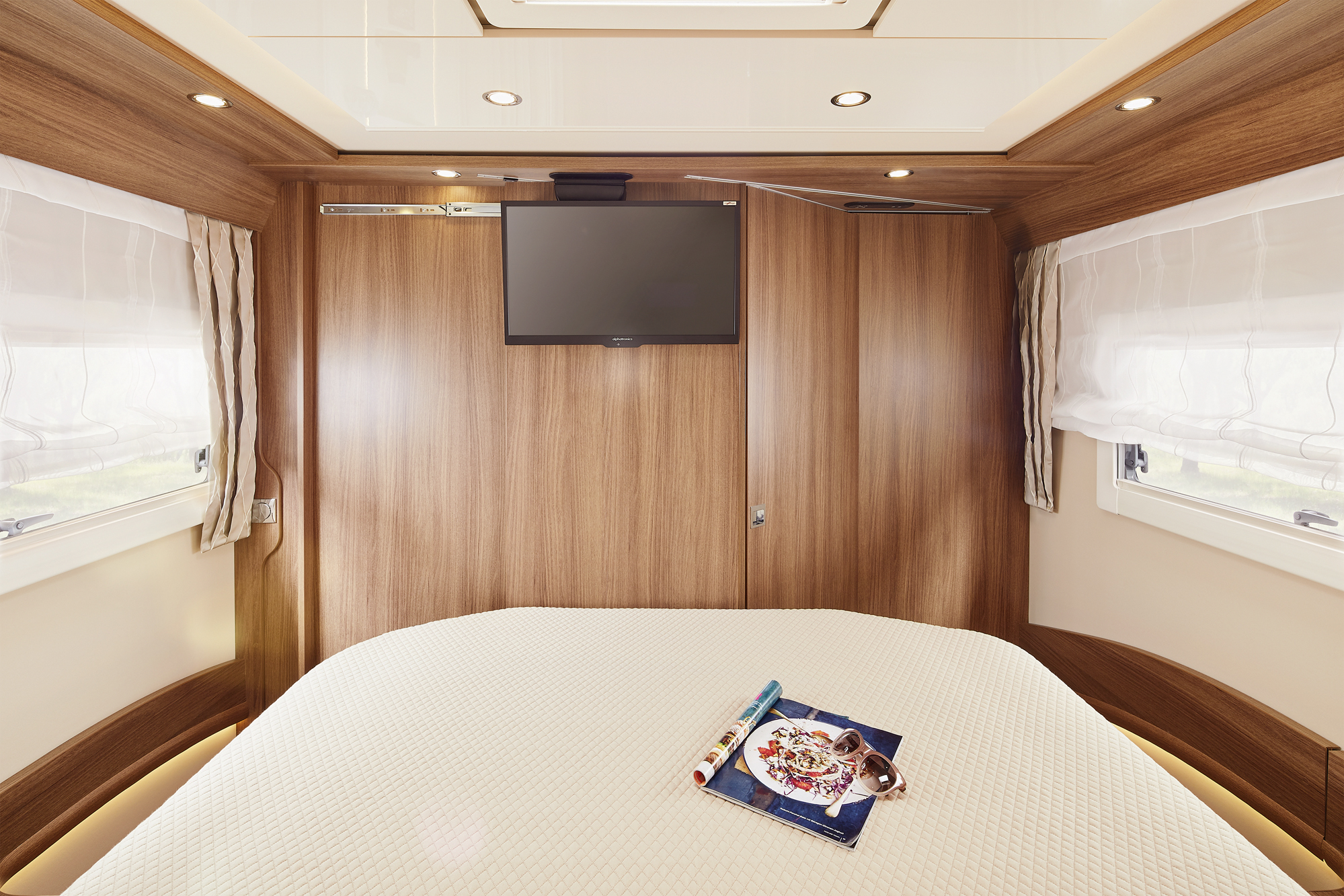 Een gezellige slaapkamer met houten wanden, een gemonteerde flatscreentelevisie en een groot bed bedekt met een witte structuurdeken. Op het bed liggen een tijdschrift, een reisgids en een opgerolde kaart. Natuurlijk licht valt binnen door twee ramen.