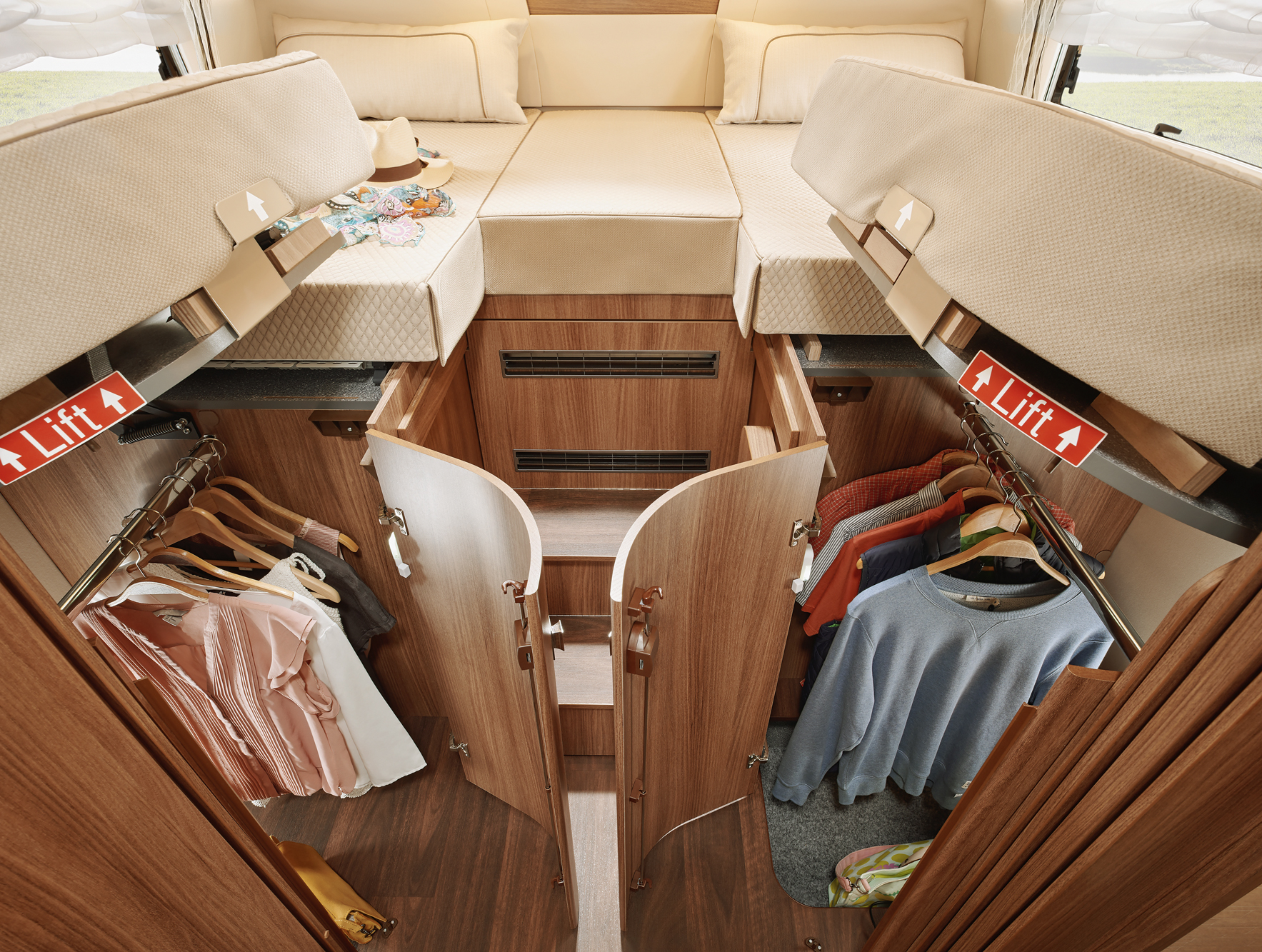 Blick in ein kompaktes Wohnmobil-Schlafzimmer mit erhöhtem Bett, zwei offenen Kleiderschränken darunter, Hängekleidung und Regalen. Hebeschilder zeigen an, dass das Bett für zusätzlichen Stauraum hochgeklappt werden kann.
