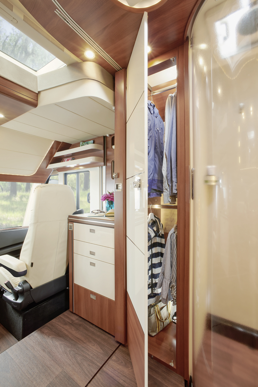 Innenraum eines modernen Wohnmobils mit einem Fahrersitz, einer kompakten Küche und einem offenen Kleiderschrank mit hängenden Kleidungsstücken; Sonnenlicht fällt durch Fenster und ein Dachfenster ein und schafft eine helle, gemütliche Atmosphäre.