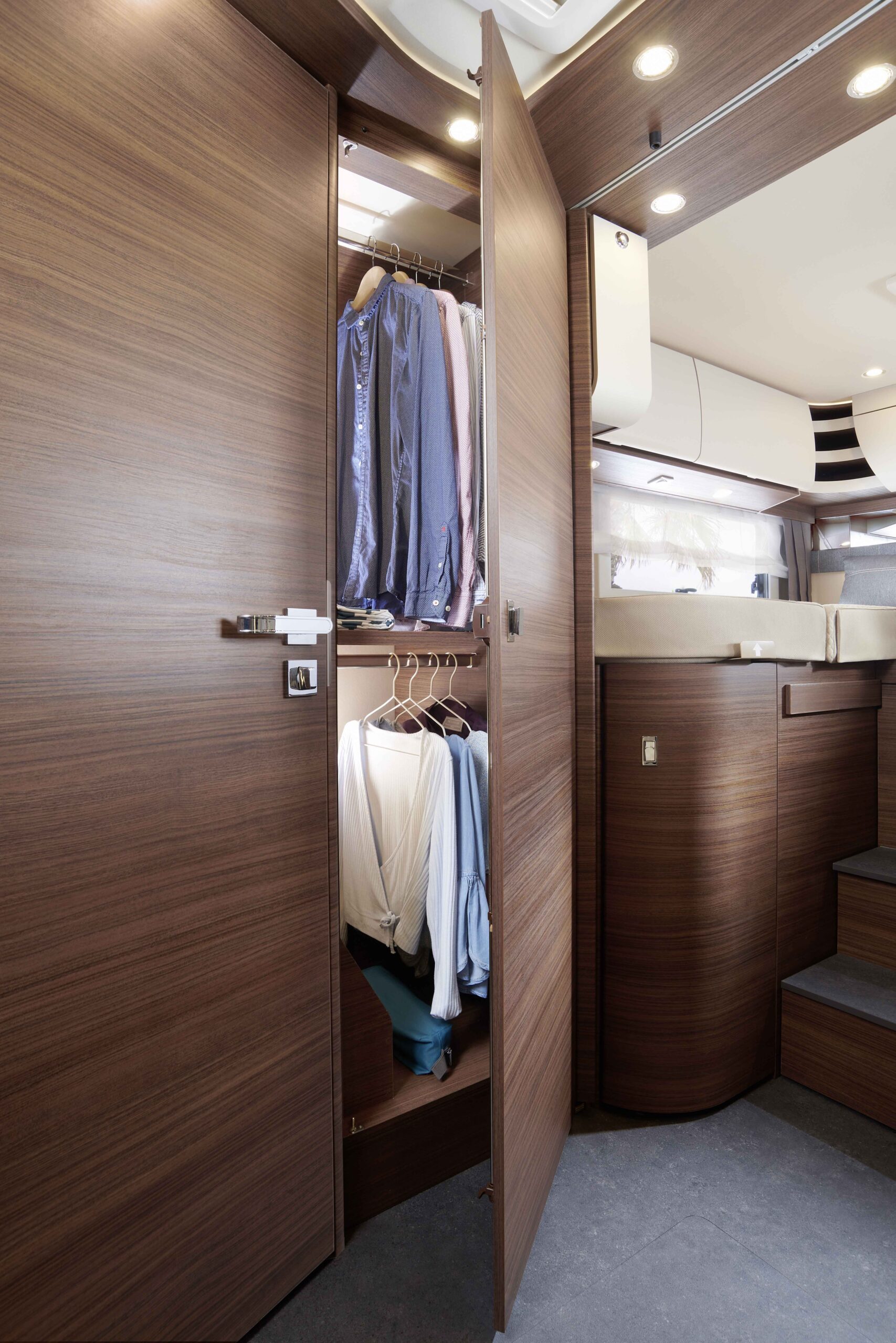Offener Holzschrank in einem modernen Wohnmobil, in dem die Kleidung ordentlich auf Bügeln hängt. Die Umgebung ist holzgetäfelt, sanft beleuchtet und bietet einen Teilblick auf die Treppe und den Wohnraum.