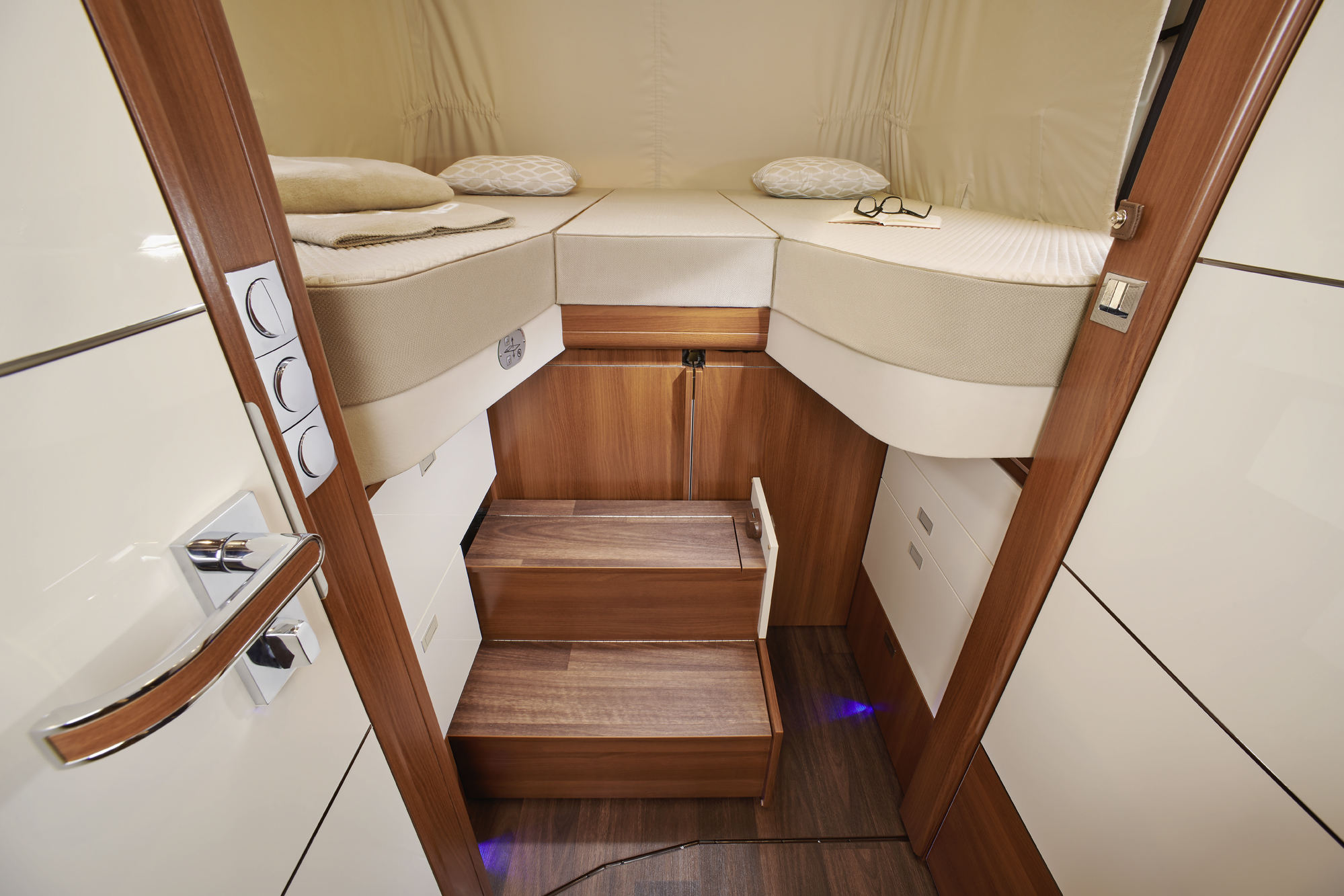 Ein modernes Wohnmobil-Schlafzimmer mit einem erhöhten Eckbett, einer Holztreppe, die zu ihm hinaufführt, sanfter Beleuchtung, Kissen und einer Brille auf dem Bett. Das Zimmer ist in Weiß und Holz gehalten.