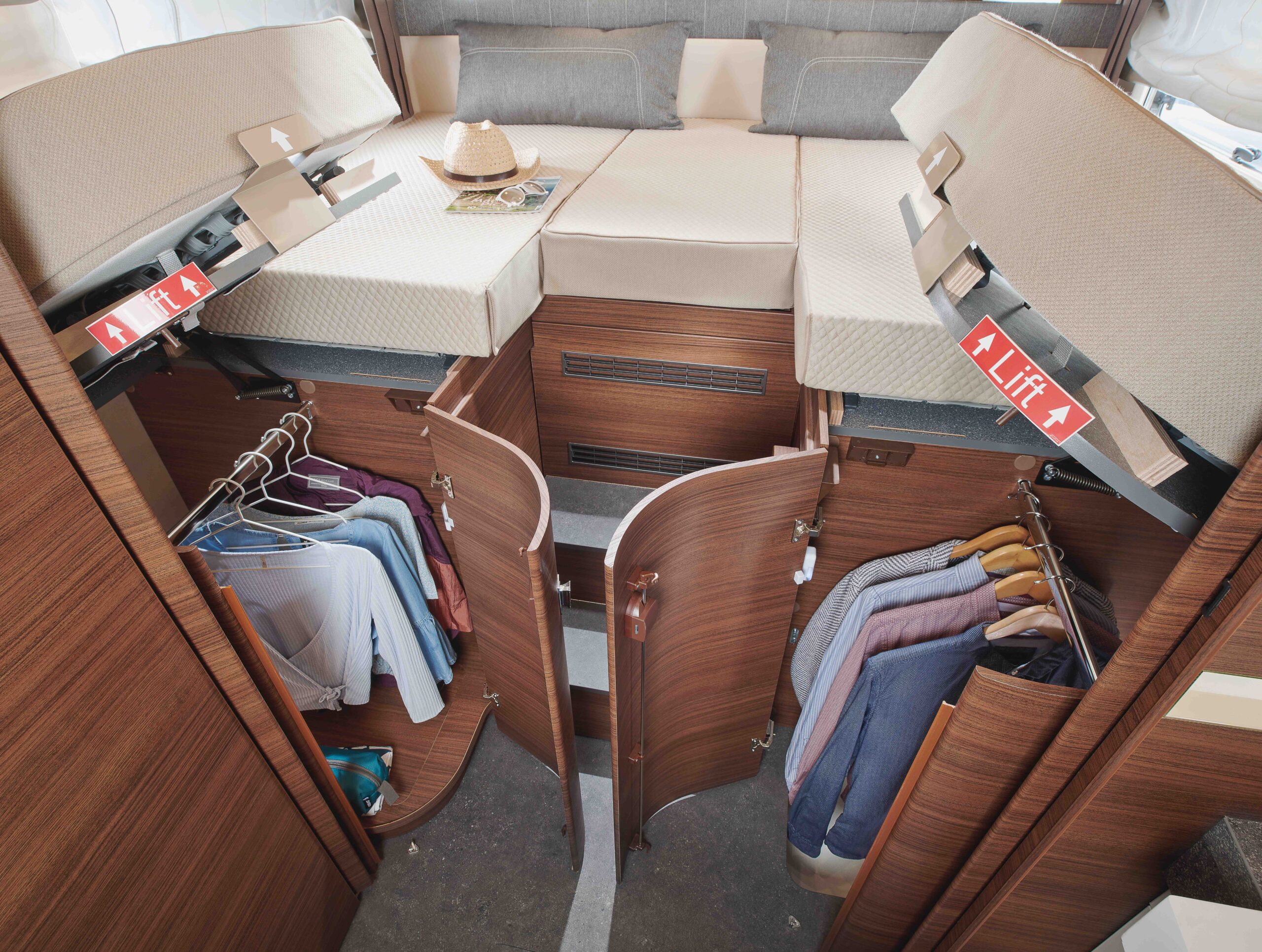 Ein ordentlich geordneter hölzerner Kleiderschrank mit offenen Türen unter einem erhöhten Bett, mit hängenden Kleidern, Regalen und zwei Fächern mit der Aufschrift "Lift" auf jeder Seite des Bettes. Ein Hut und ein Buch liegen auf dem Bett.