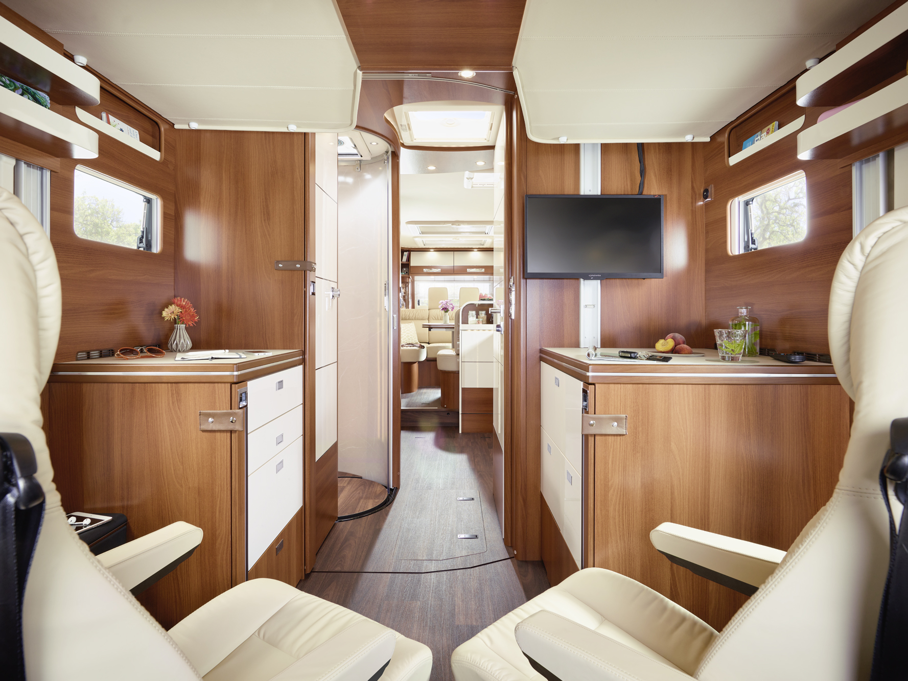 Innenraum eines modernen Wohnmobils mit Holzvertäfelung, zwei cremefarbenen Sitzen, einem kleinen Schreibtisch mit einem Fernseher, Schränken und großen Fenstern. Im Hintergrund sind eine Küche und ein Essbereich zu sehen. Blumen und Früchte schmücken den Raum.