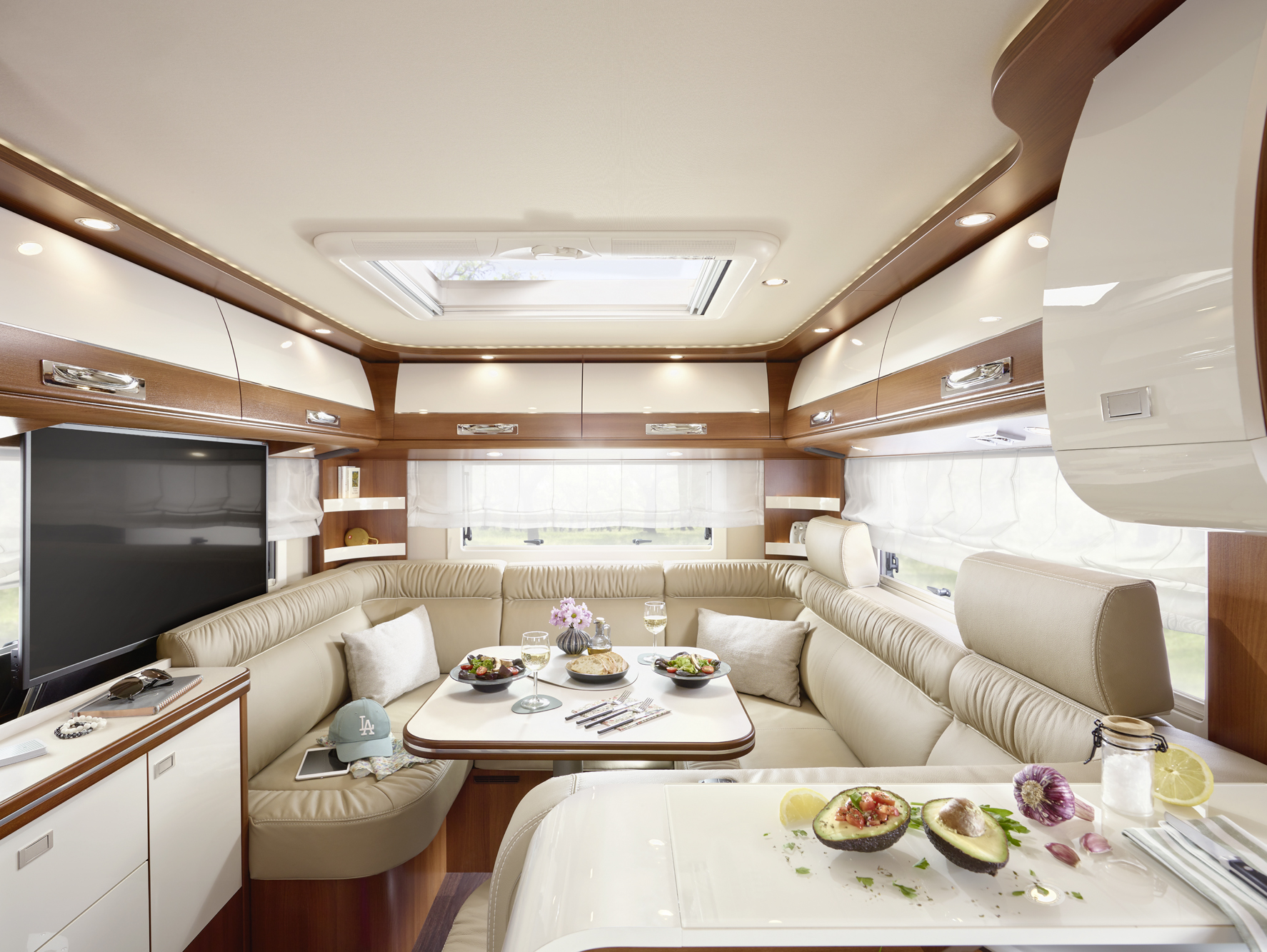 Das Foto zeigt ein modernes Wohnmobil-Interieur mit beigen Ledersitzen, einem Esstisch mit Speisen und Getränken, Holzschränken, einem Fernseher und einem Dachfenster, das eine helle, gemütliche Atmosphäre schafft.