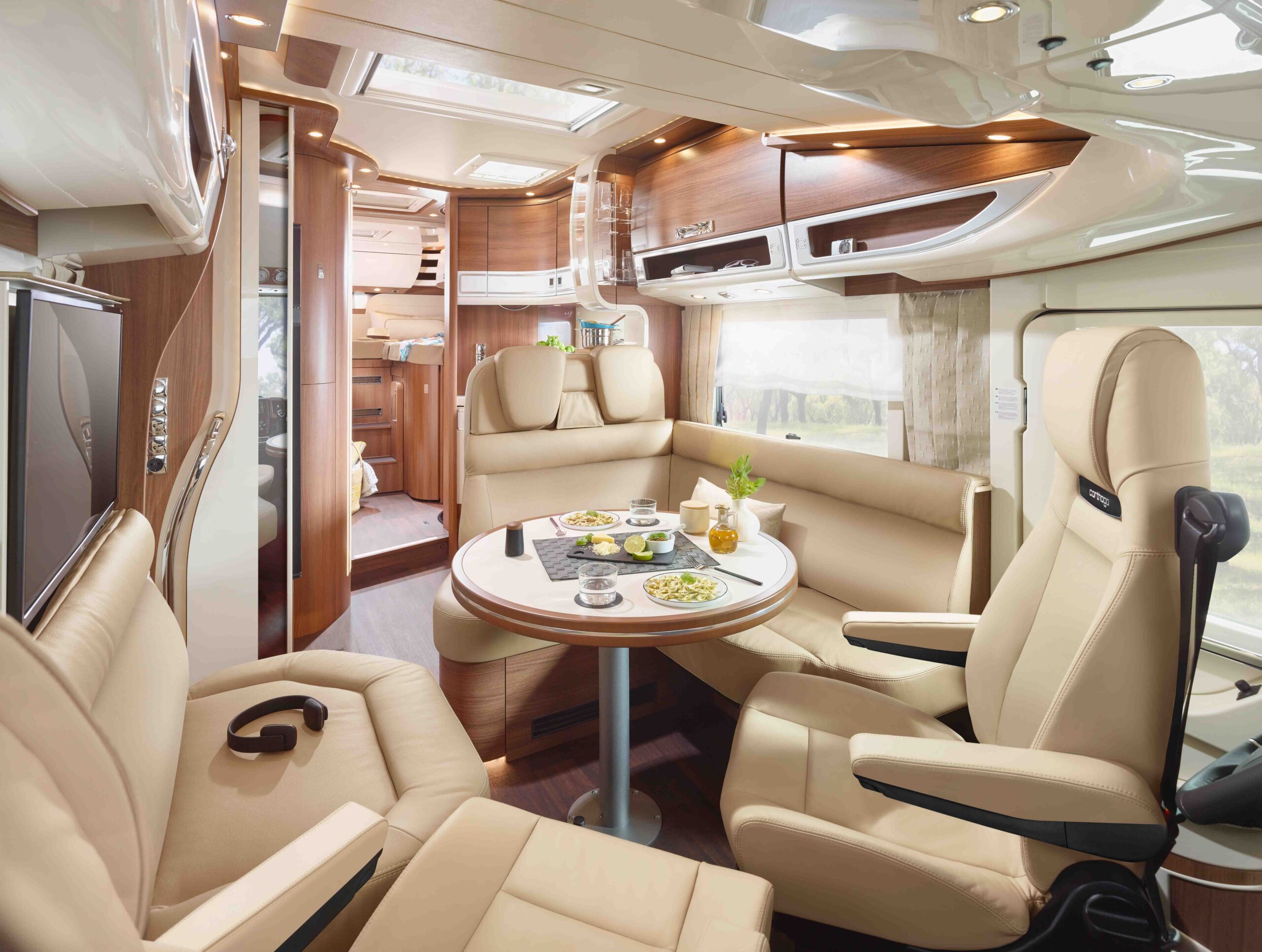 Der Innenraum eines luxuriösen Wohnmobils mit cremefarbenen Ledersitzen, die um einen runden Esstisch mit Tellern, Tassen und Snacks angeordnet sind. Der Raum wirkt modern, ist gut beleuchtet und verfügt über Holzakzente und eingebaute Stauräume.