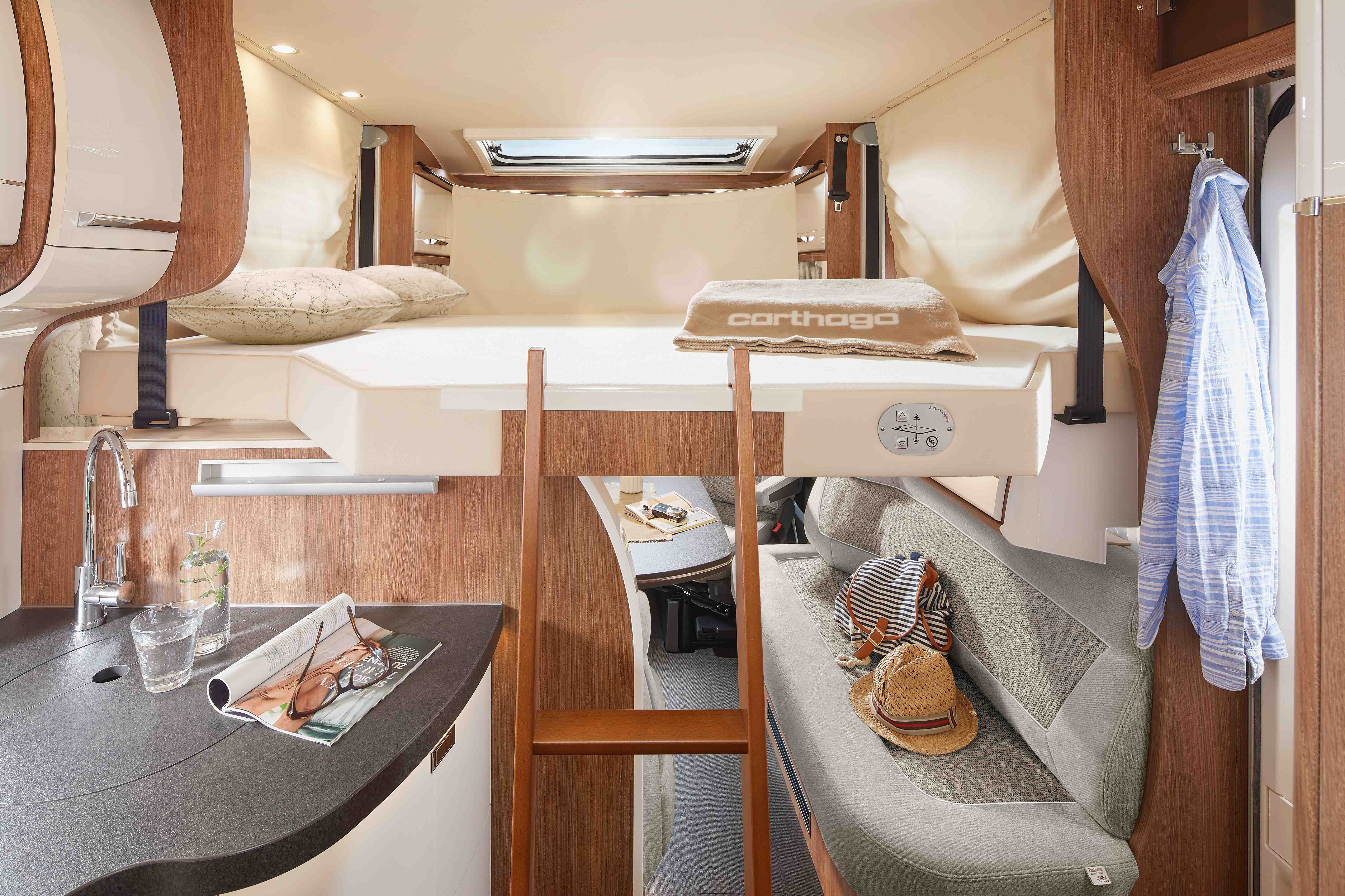 Intérieur d'un camping-car comprenant un lit en mezzanine avec une serviette et un oreiller, une échelle en bois, un petit coin cuisine avec un évier et un verre, et un coin salon avec un chapeau et un sac. Éclairage chaleureux et touches de bois dans tout le véhicule.