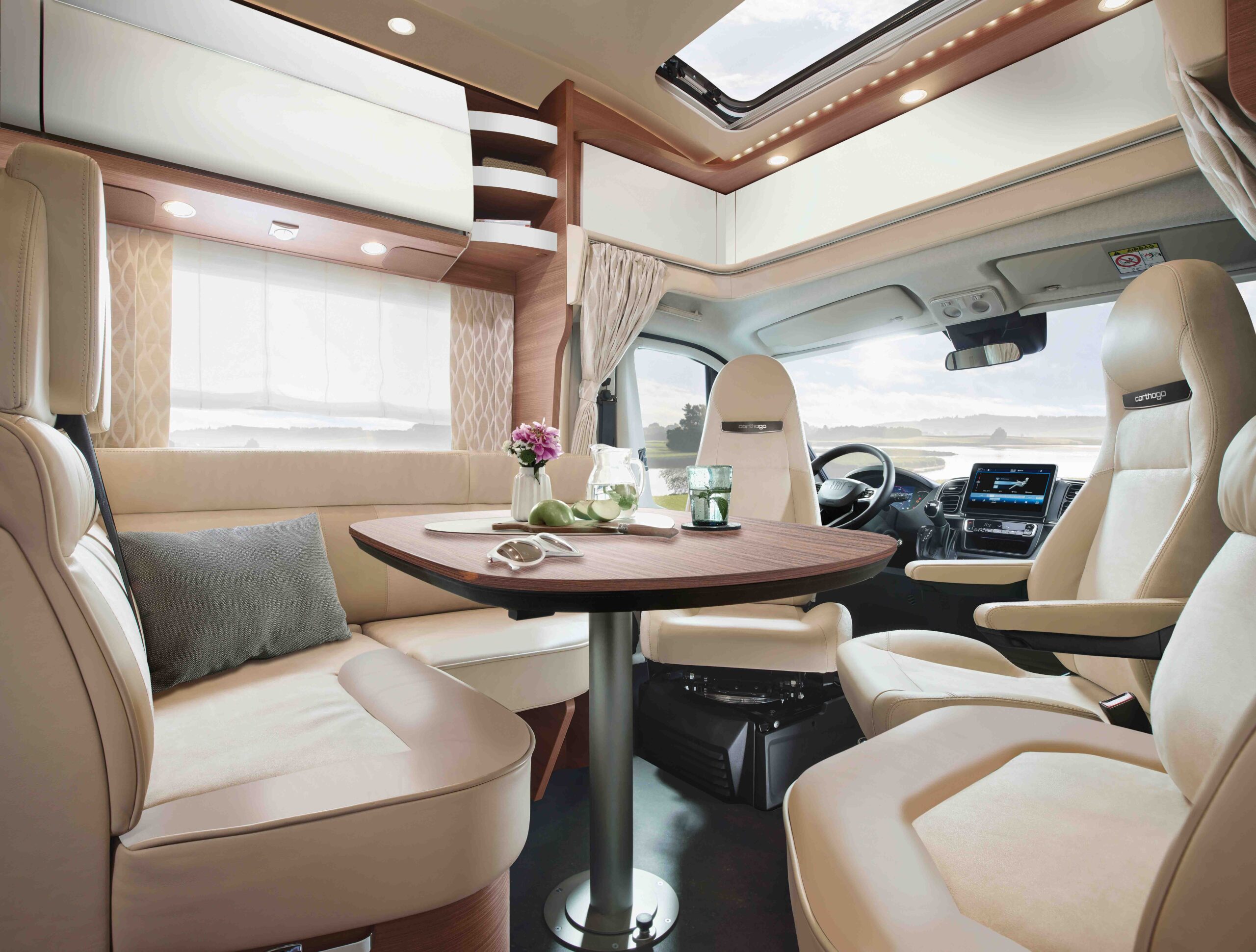 Modernes Wohnmobil-Interieur mit cremefarbenen Ledersitzen, einem runden Esstisch mit Geschirr und Blumen, großen Fenstern mit Vorhängen und einem Dachfenster, die eine helle und gemütliche Atmosphäre schaffen. Der Fahrerbereich ist im Hintergrund zu sehen.