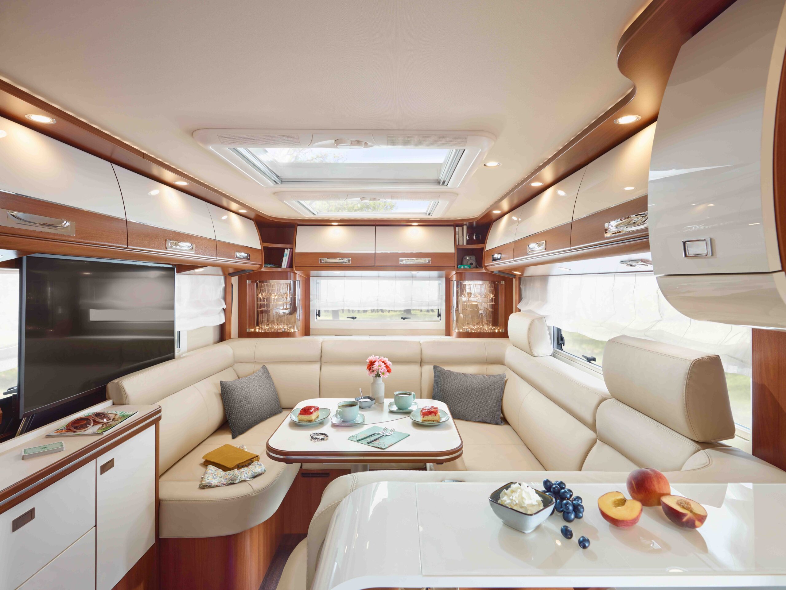 Modernes Wohnmobil-Interieur mit cremefarbenen Sitzgelegenheiten, einem weißen Frühstückstisch, Obst auf der Theke, Holzschränken, einem großen Fenster, Oberlichtern und einem Dachfenster, die eine helle, gemütliche Atmosphäre schaffen.