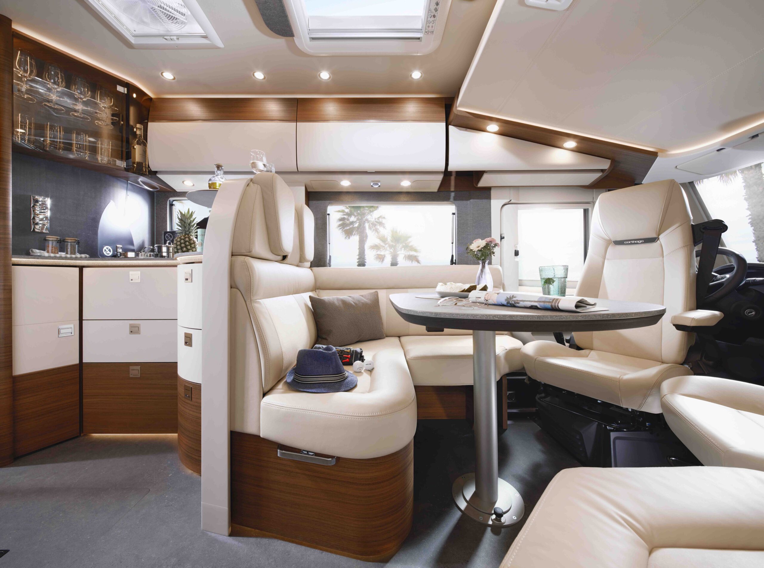 Een modern interieur met beige lederen zetels, een ronde eettafel, ingebouwde kasten, glaswerk en grote ramen die zonlicht binnenlaten. Een hoed, zonnebril en drankjes staan op de tafel en stoelen.