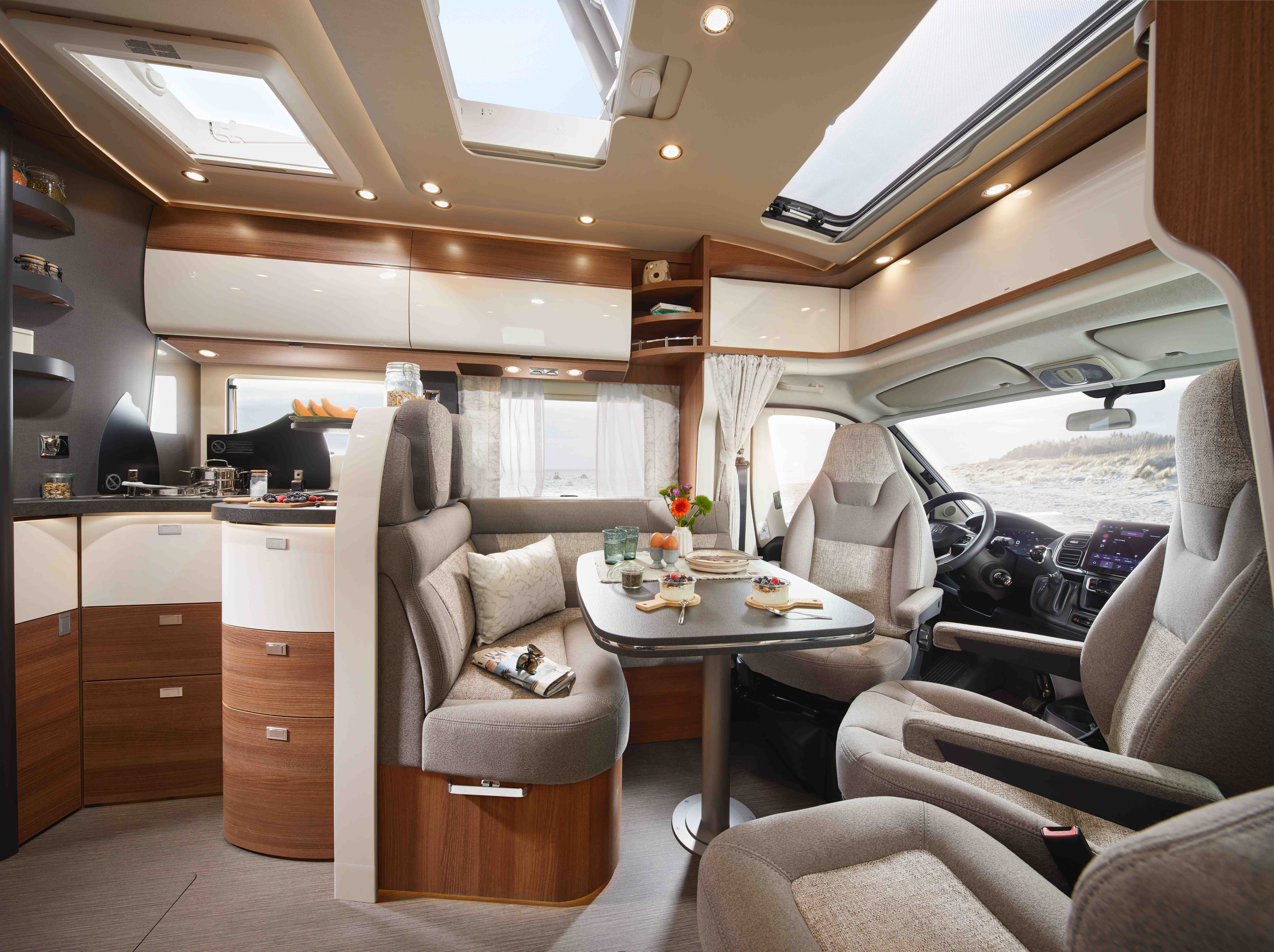Intérieur moderne d'un camping-car avec des sièges confortables, une table à manger pour un repas, un coin cuisine avec des ustensiles, de grandes fenêtres, des puits de lumière et des armoires en bois clair. À l'extérieur, un paysage de plage sablonneuse est visible à travers les fenêtres.