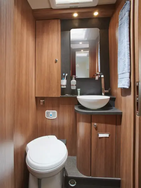Kompaktes Badezimmer in einem Carthago C2-Tourer mit Holzschränken, einer weißen Toilette, einem modernen Waschbecken mit geschwungenem Wasserhahn, einem Wandspiegel, Zahnbürsten in Halterungen, einem kleinen Schlauch und einem blauen Handtuch an der rechten Wand. Helle Deckenbeleuchtung.