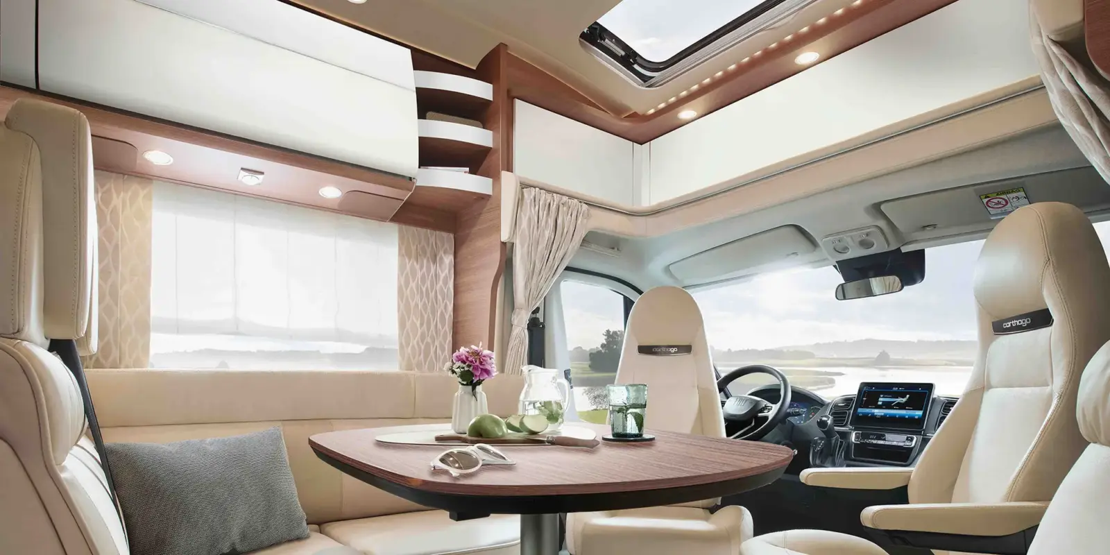 Der Innenraum eines Carthago Chic C Line T Wohnmobils ist mit cremefarbenen Sitzen, einem runden Holztisch mit Blumen und Snacks, großen Fenstern mit Vorhängen und einem Schiebedach ausgestattet und schafft so eine helle, gemütliche Atmosphäre mit Blick auf die Landschaft.