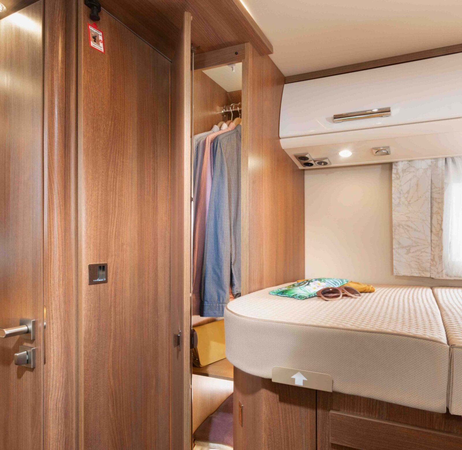 Holzgetäfelter Schrank mit Kleiderbügeln und Regalen im Inneren, daneben ein Bett mit einem Buch und einer Brille darauf, in einem kompakten, gemütlichen Carthago C Tourer teilintegrierten Wohnraum mit warmer Beleuchtung.