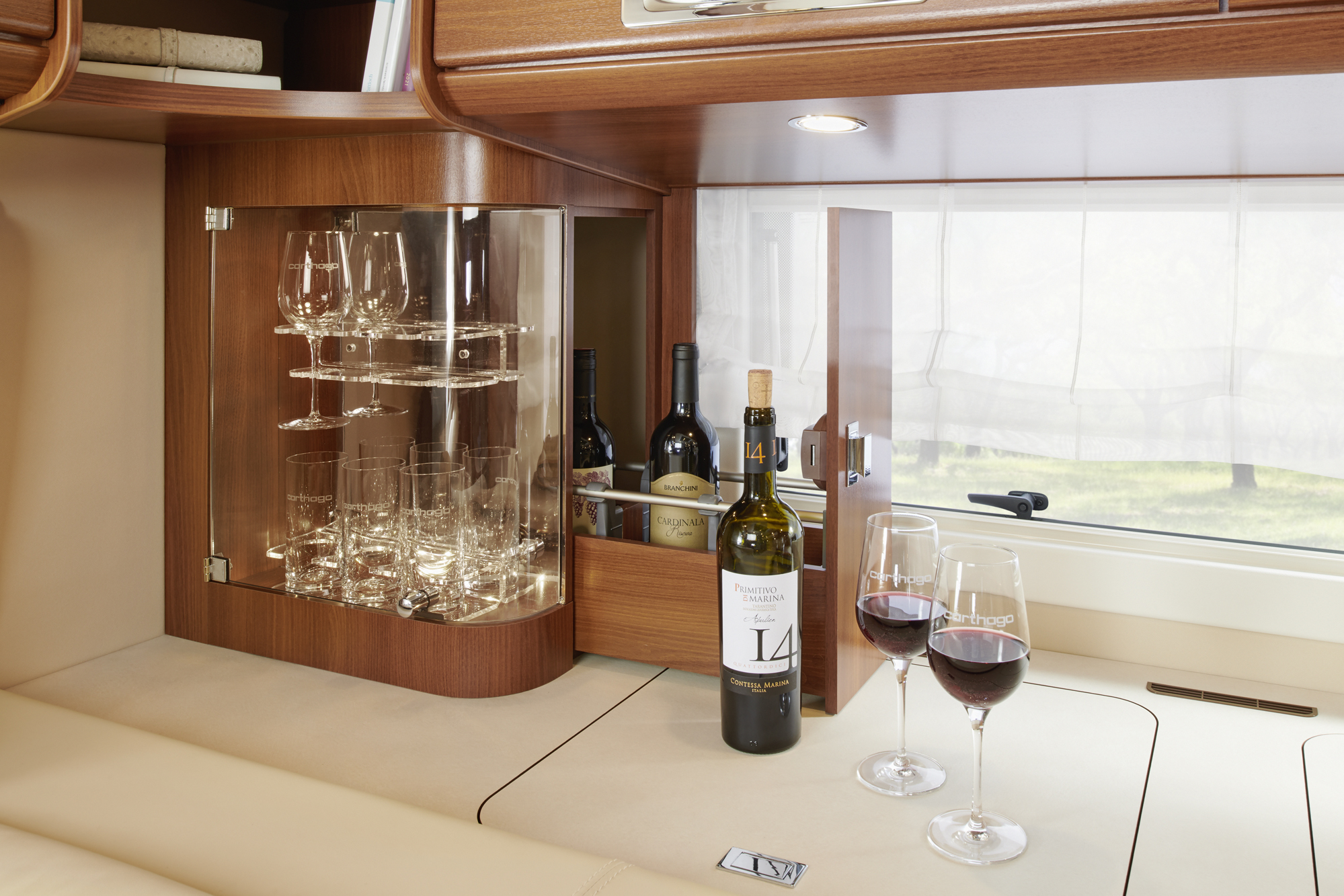 Ein moderner Holzschrank mit Gläsern, Weinflaschen und zwei Gläsern Rotwein auf einer Arbeitsplatte neben einem Fenster mit durchsichtigen Vorhängen.