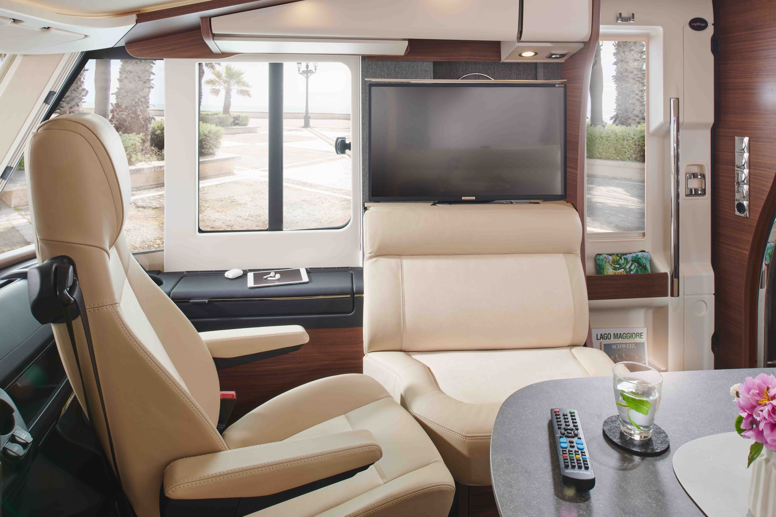 Het interieur van een moderne camper met beige lederen zetels, een gemonteerde tv, een tafel met drankjes en afstandsbedieningen, en grote ramen met palmbomen en een zonnig buitendecor.