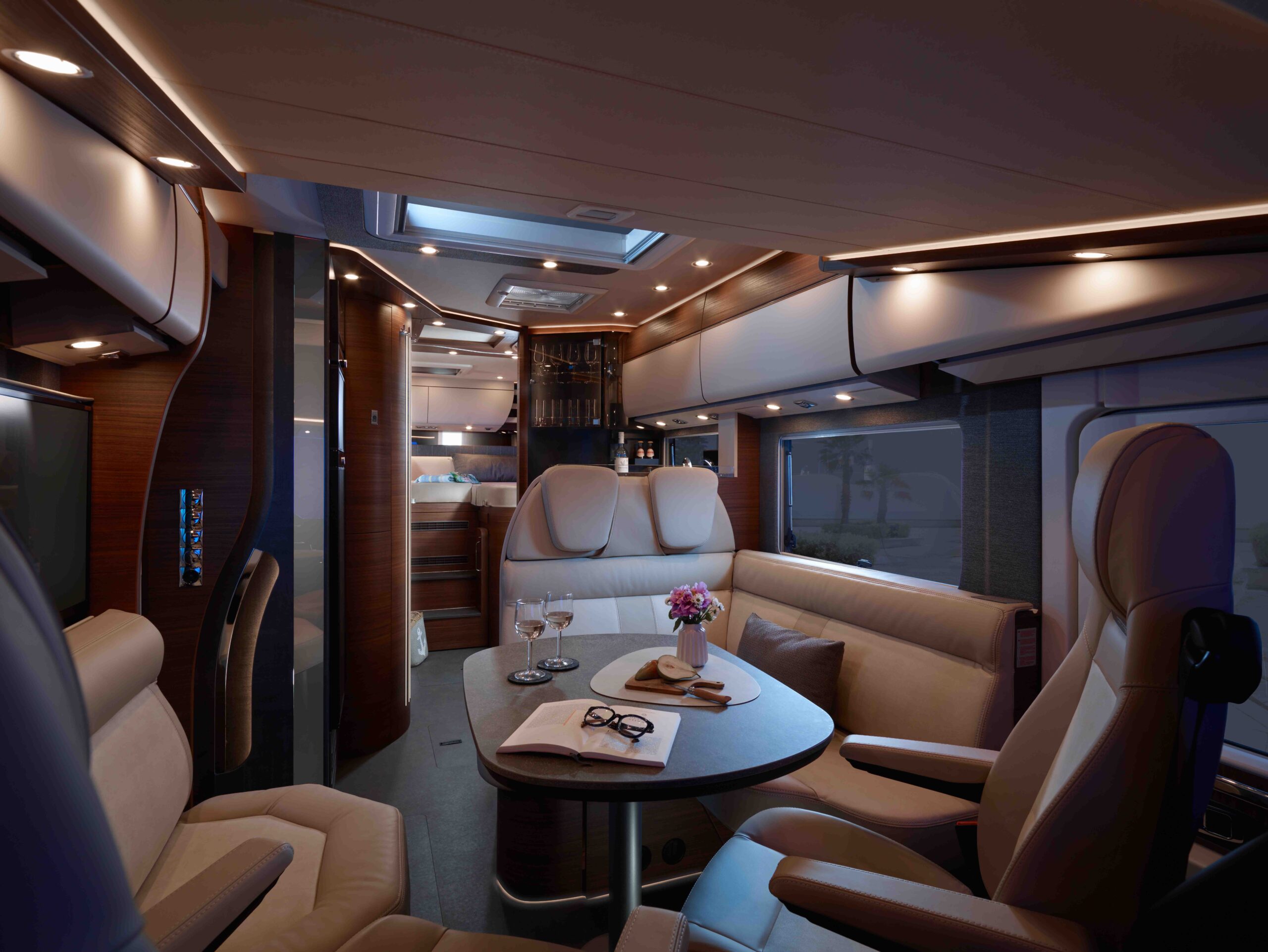 Luxuriöses Wohnmobil-Interieur mit Ledersitzen, einem runden Esstisch mit Weingläsern, einem Buch und Blumen. Sanfte Umgebungsbeleuchtung unterstreicht die dunkle Holzverkleidung und das moderne Design. Im Hintergrund ist ein Bett zu sehen.