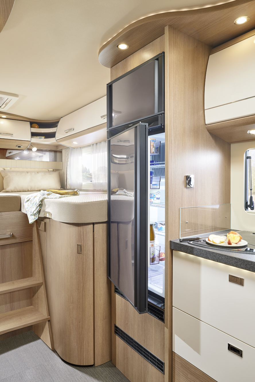Intérieur d'un camping-car moderne comprenant une cuisine compacte avec un comptoir, un réfrigérateur dont la porte est ouverte, des armoires en bois, des marches menant à un lit surélevé et de la lumière naturelle provenant d'une fenêtre.