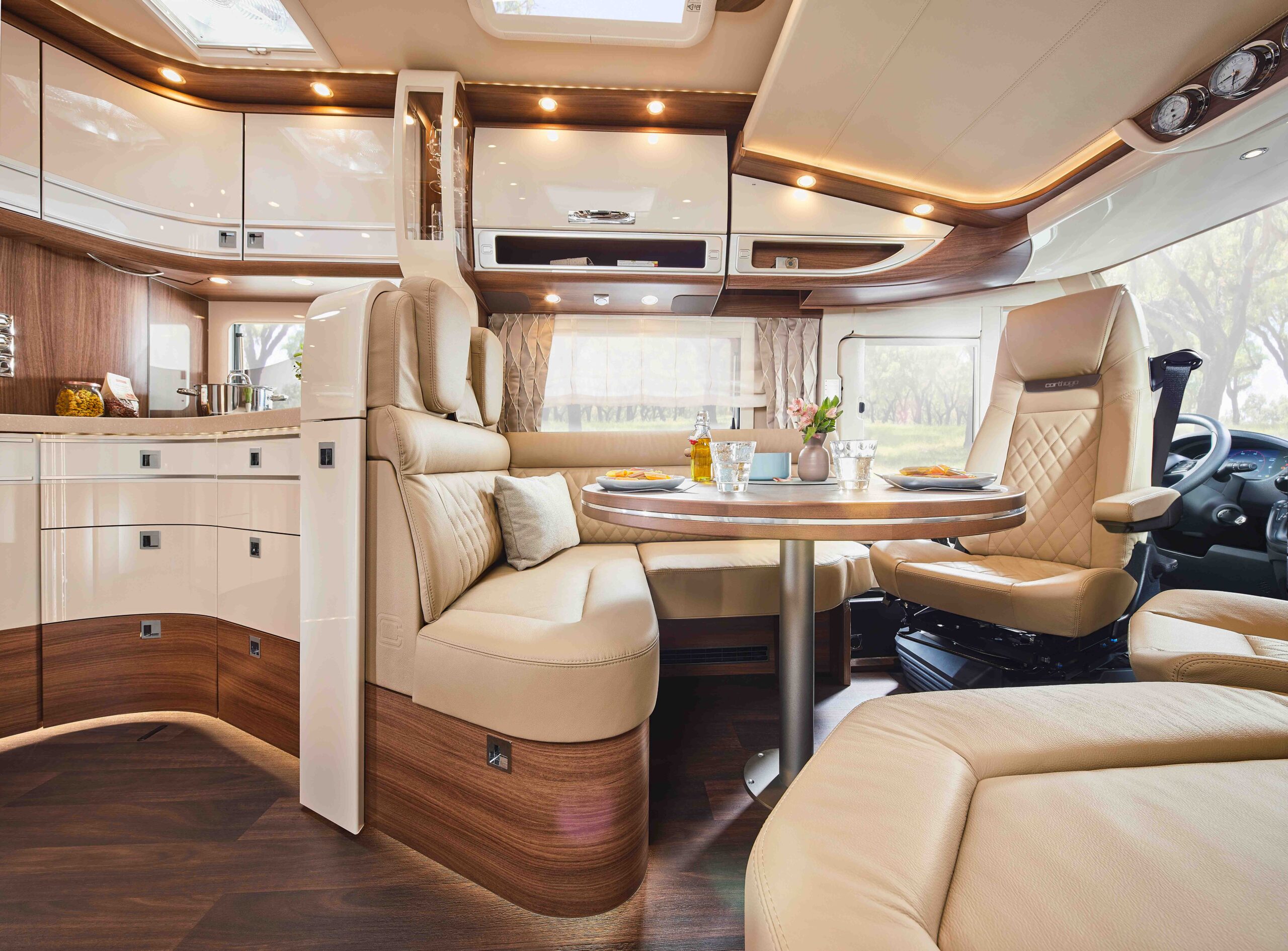 Ein luxuriöses Wohnmobil-Interieur mit cremefarbenen Ledersitzen, einem runden Esstisch mit Frühstückszubehör, glänzenden Schränken, Holzakzenten und großen Fenstern, die natürliches Licht hereinlassen.