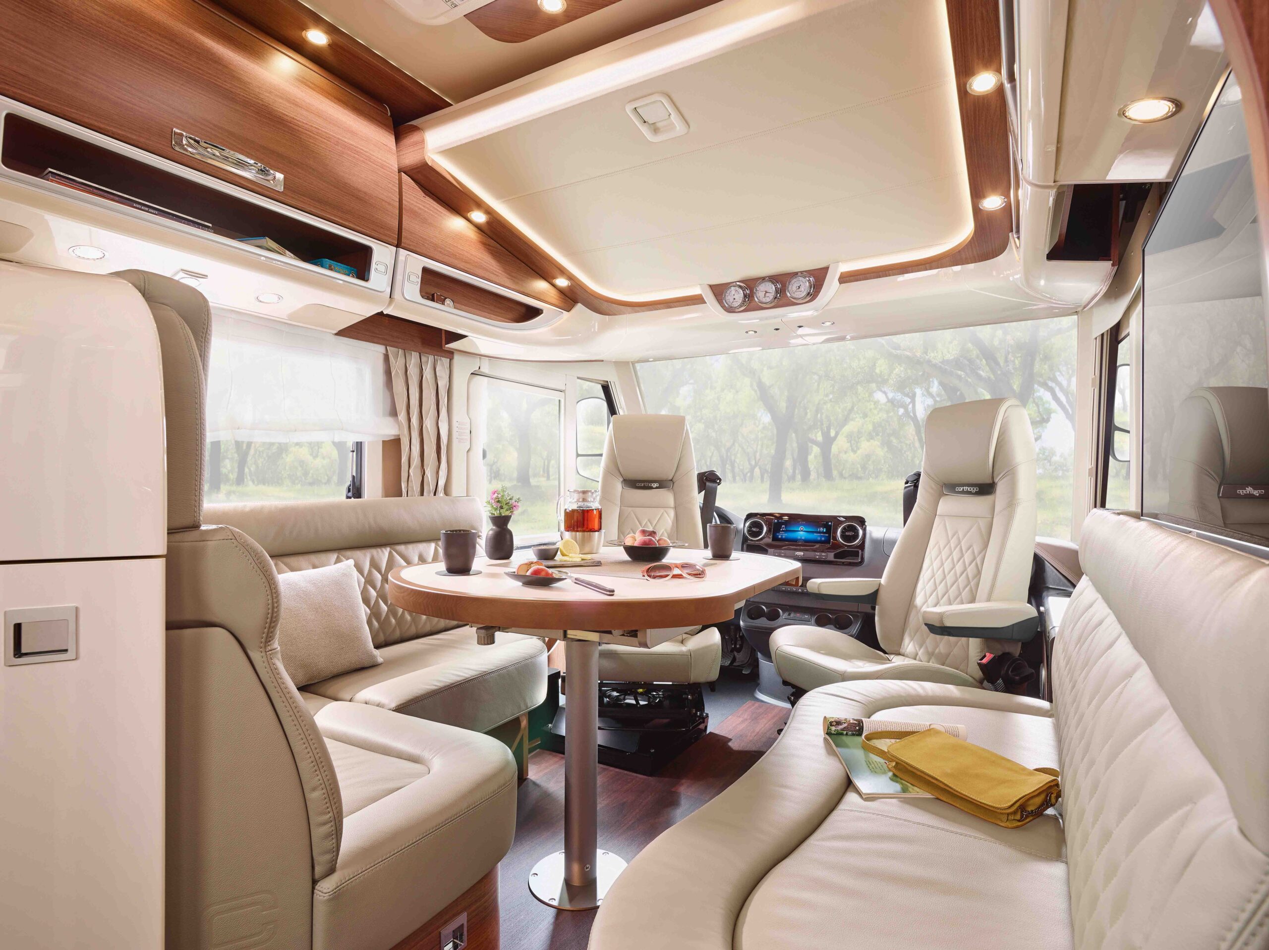 Ein luxuriöses Wohnmobil-Interieur mit cremefarbenen Ledersitzen, einem runden Holztisch, großen Fenstern und moderner Beleuchtung. Auf dem Tisch stehen Getränke und Snacks, während im Hintergrund die Küche und die Schränke zu sehen sind.