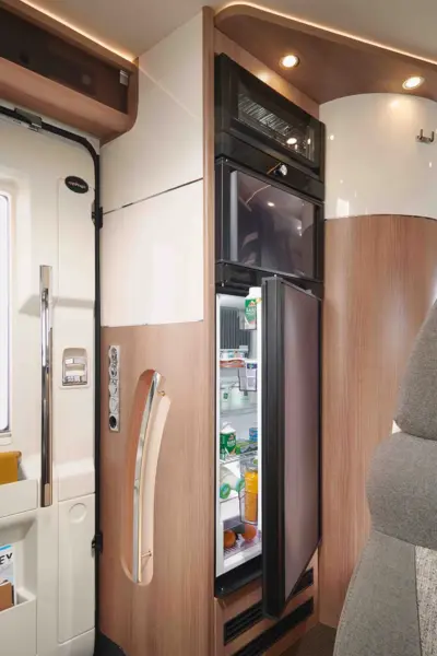 Die kompakte Wohnmobilküche im Carthago C Tourer verfügt über einen hohen, modernen Kühlschrank mit offener Tür, in dem die Lebensmittel und Getränke übersichtlich angeordnet sind. Oberhalb des Kühlschranks befindet sich eine eingebaute Mikrowelle, umgeben von warmen Holzschränken.