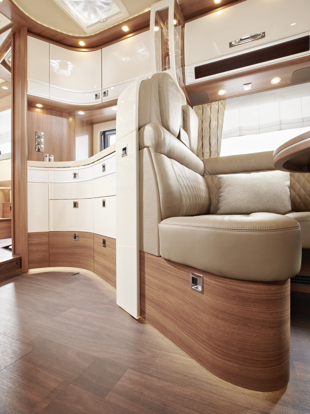 Modern RV interior featuring light brown wood flooring, curved cream and wood cabinetry, and beige leather seating. Soft lighting highlights a cozy, upscale space with ample storage and a dining area.