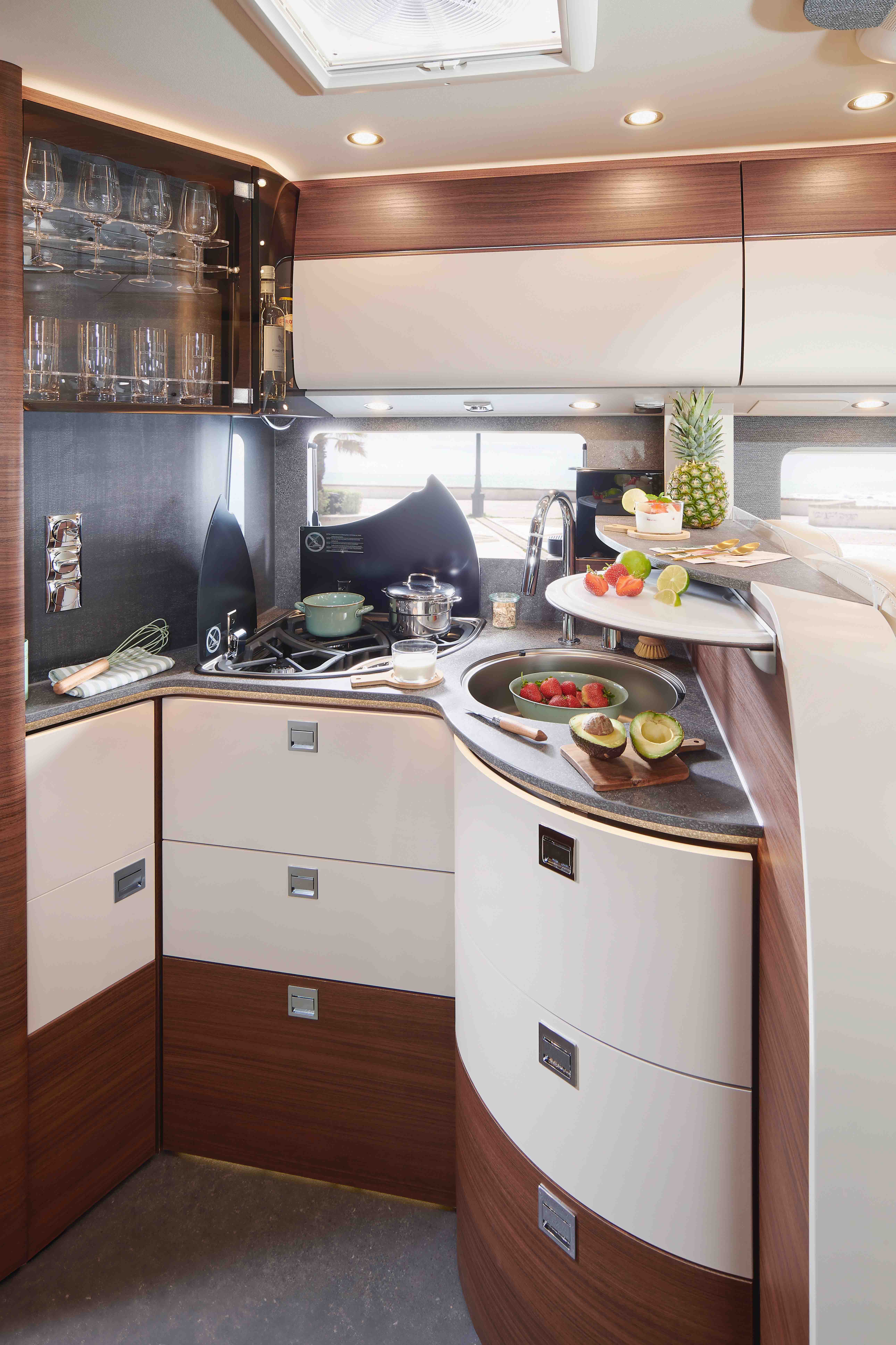 Modern RV kitchen with sleek wood and white cabinets, a small sink, stove, and countertop with fresh fruits, vegetables, and a pineapple. Glassware is stored in an overhead cabinet and natural light comes through a skylight.