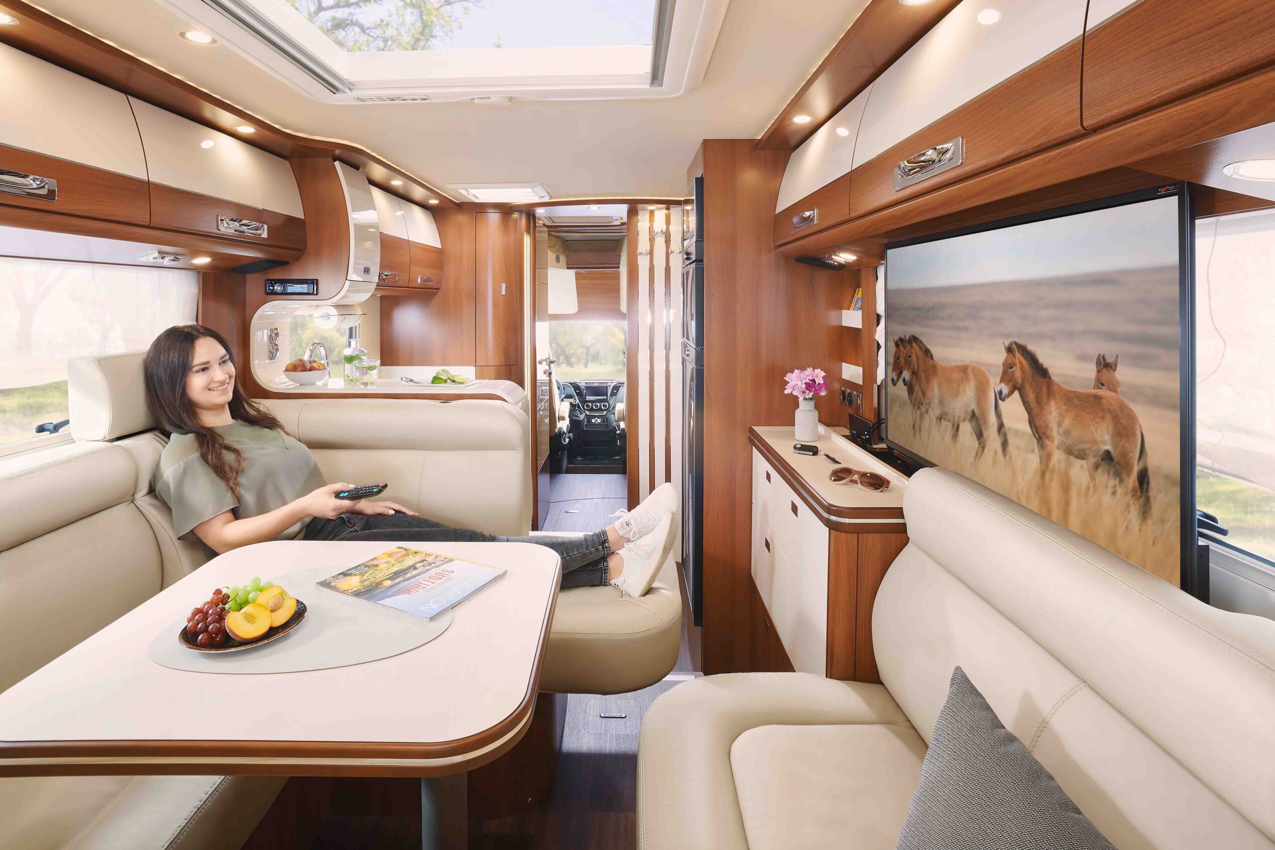 Eine Frau entspannt sich auf einem Sofa in einem modernen Wohnmobil, hält eine Fernbedienung in der Hand und beobachtet Pferde auf einem großen Fernseher. Das Innere des Wohnmobils ist mit Holzverkleidung, einem Tisch mit Obst und Zeitschriften sowie Umgebungsbeleuchtung ausgestattet.