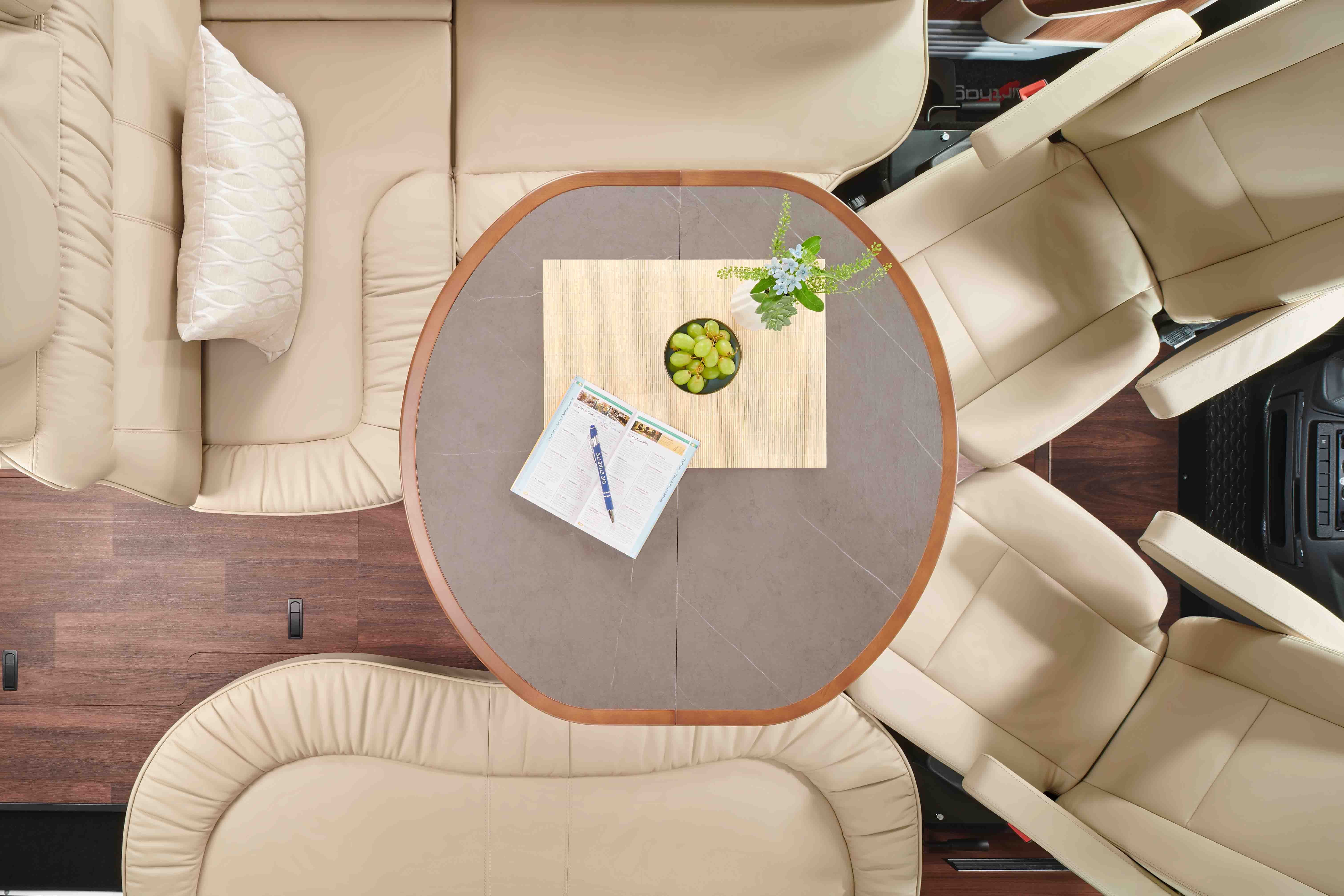 Overhead view of a luxury RV interior with plush cream leather seating and a circular table with grapes, flowers, and a magazine.