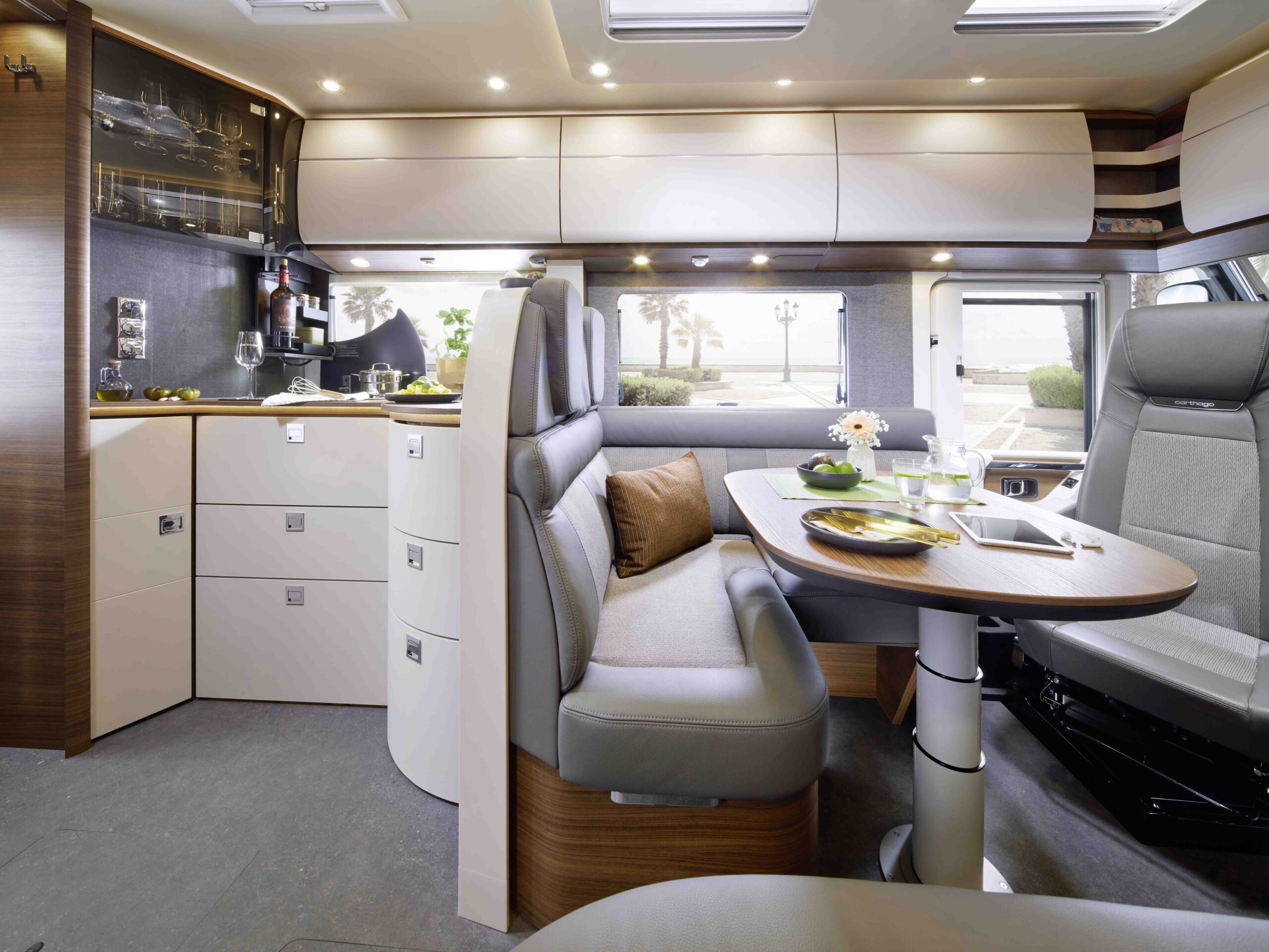 Modernes Wohnmobil-Interieur mit einer kompakten Küche, Hängeschränken und einem Essbereich mit gepolsterter Sitzbank und Tisch für zwei Personen. Große Fenster lassen natürliches Licht herein und geben den Blick auf die Natur frei.