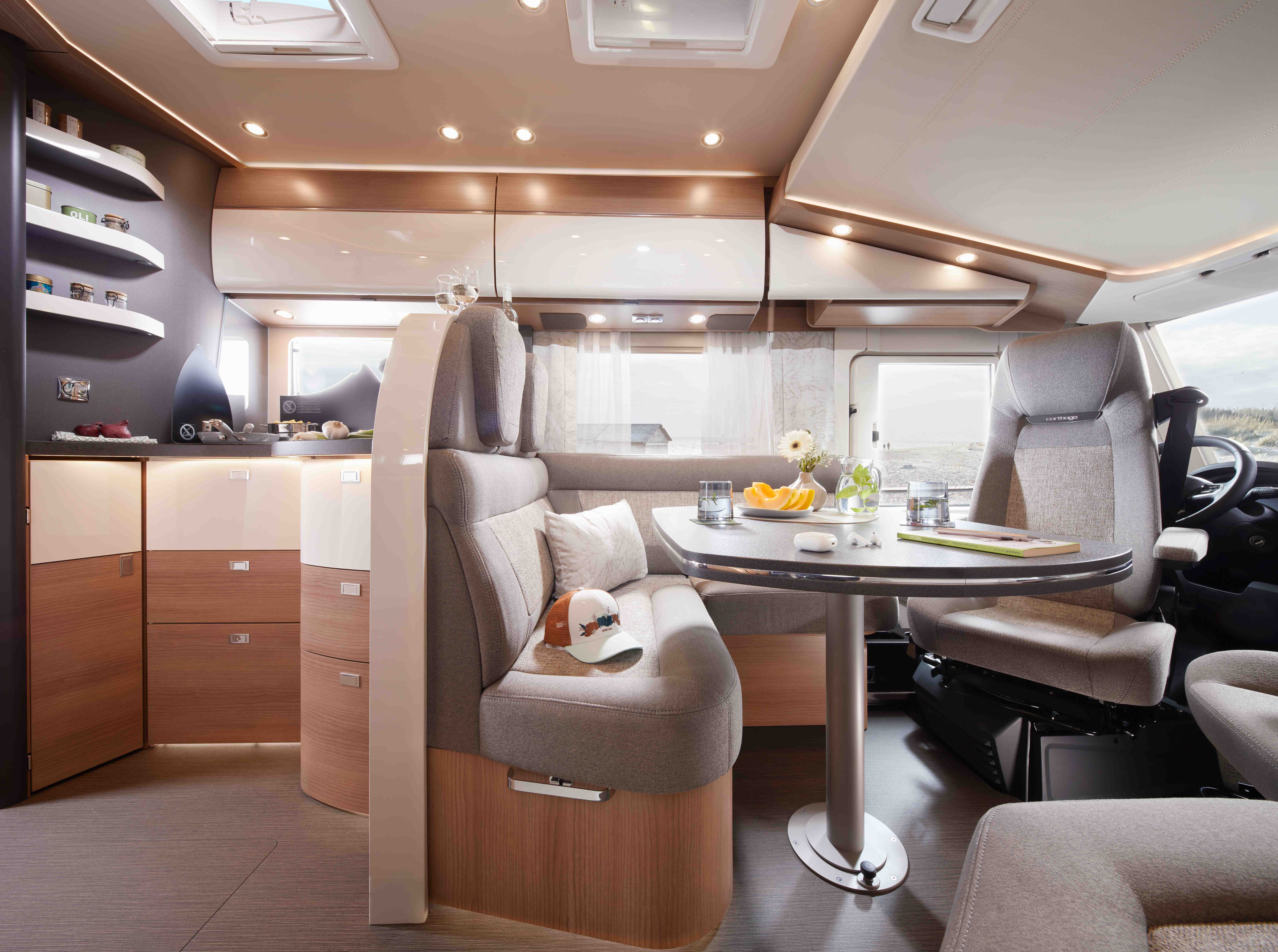 Intérieur de camping-car moderne avec des armoires en bois clair, des étagères incurvées et des sièges de couleur neutre autour d'une table garnie d'articles de petit-déjeuner et de fleurs. De grandes fenêtres laissent entrer la lumière naturelle, créant une atmosphère lumineuse et confortable.