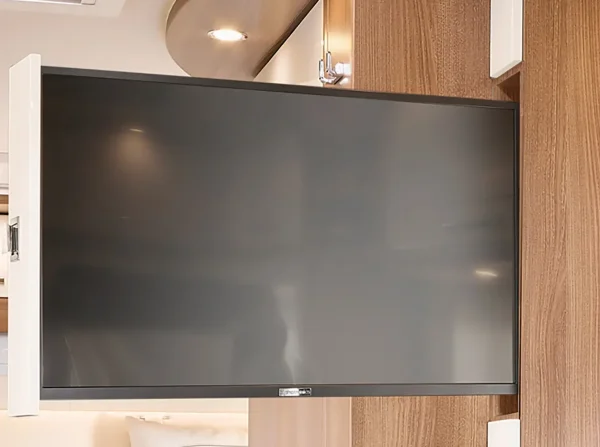 Ein Flachbildfernseher ist auf einer schwenkbaren Halterung an einer Holzwand im Inneren eines modernen Carthago C Tourer Teilintegrierten Wohnmobils angebracht. Der Fernseher ist nach außen gedreht und bringt die hellen Holzakzente und das neutrale Dekor des Raums zur Geltung.