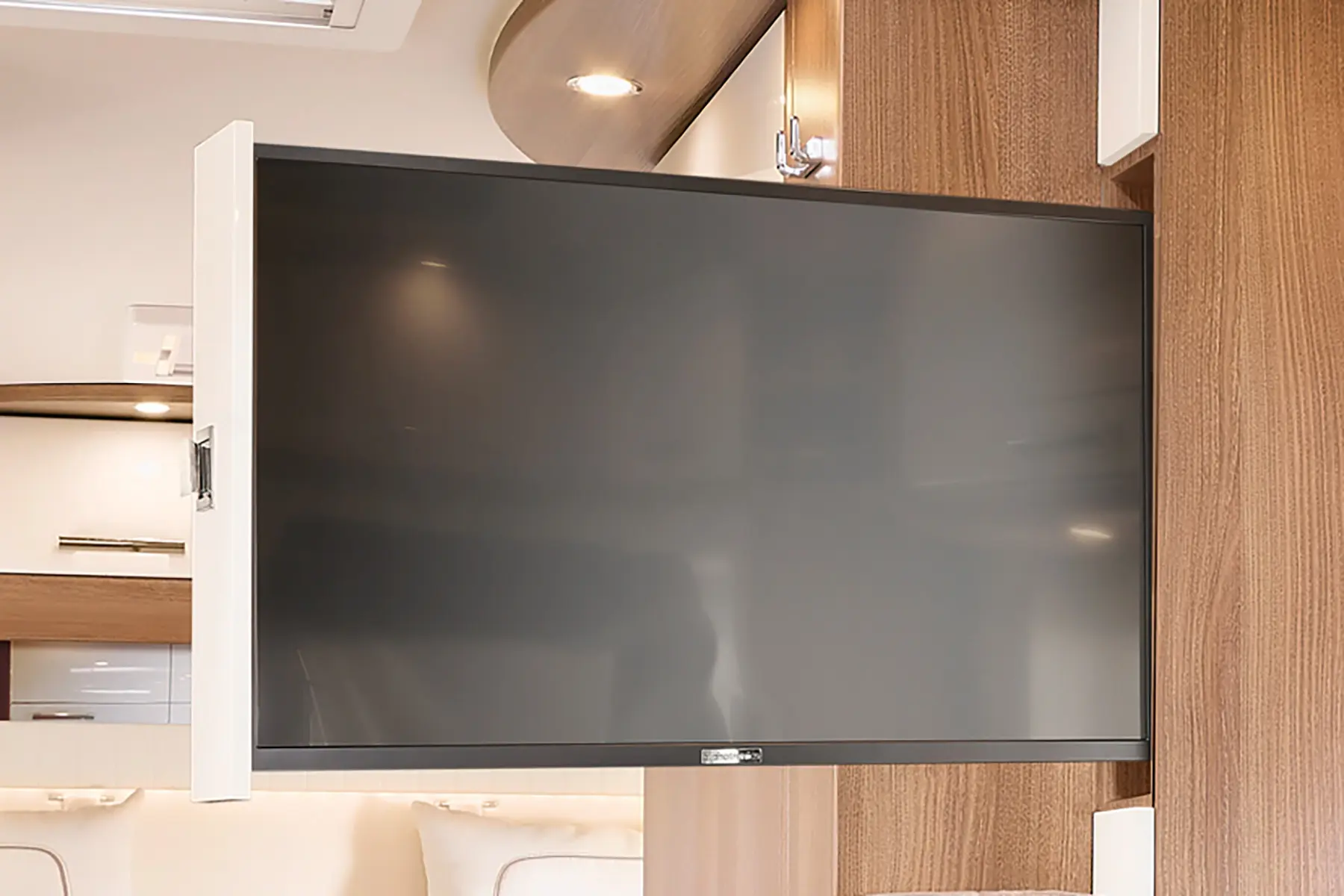 Ein Flachbildfernseher ist auf einer schwenkbaren Halterung an einer Holzwand im Inneren eines modernen Carthago C Tourer Teilintegrierten Wohnmobils angebracht. Der Fernseher ist nach außen gedreht und bringt die hellen Holzakzente und das neutrale Dekor des Raums zur Geltung.