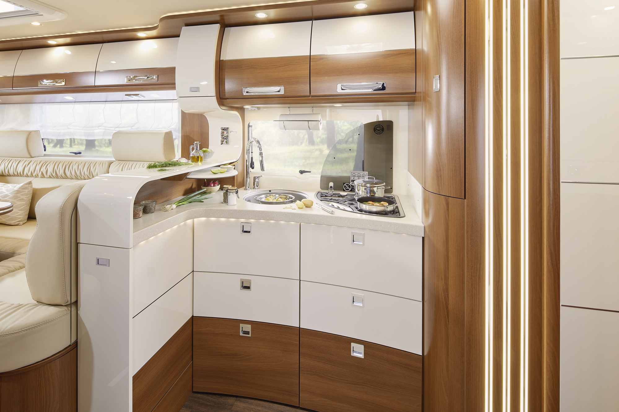 Moderne Wohnmobilküche mit glänzend weißen und hölzernen Schränken, eingebautem Herd, Spüle und Arbeitsplatte. Es gibt Teller mit Essen, Utensilien und natürliches Licht, das durch ein großes Fenster einfällt. Links ist ein gemütlicher Sitzbereich zu sehen.