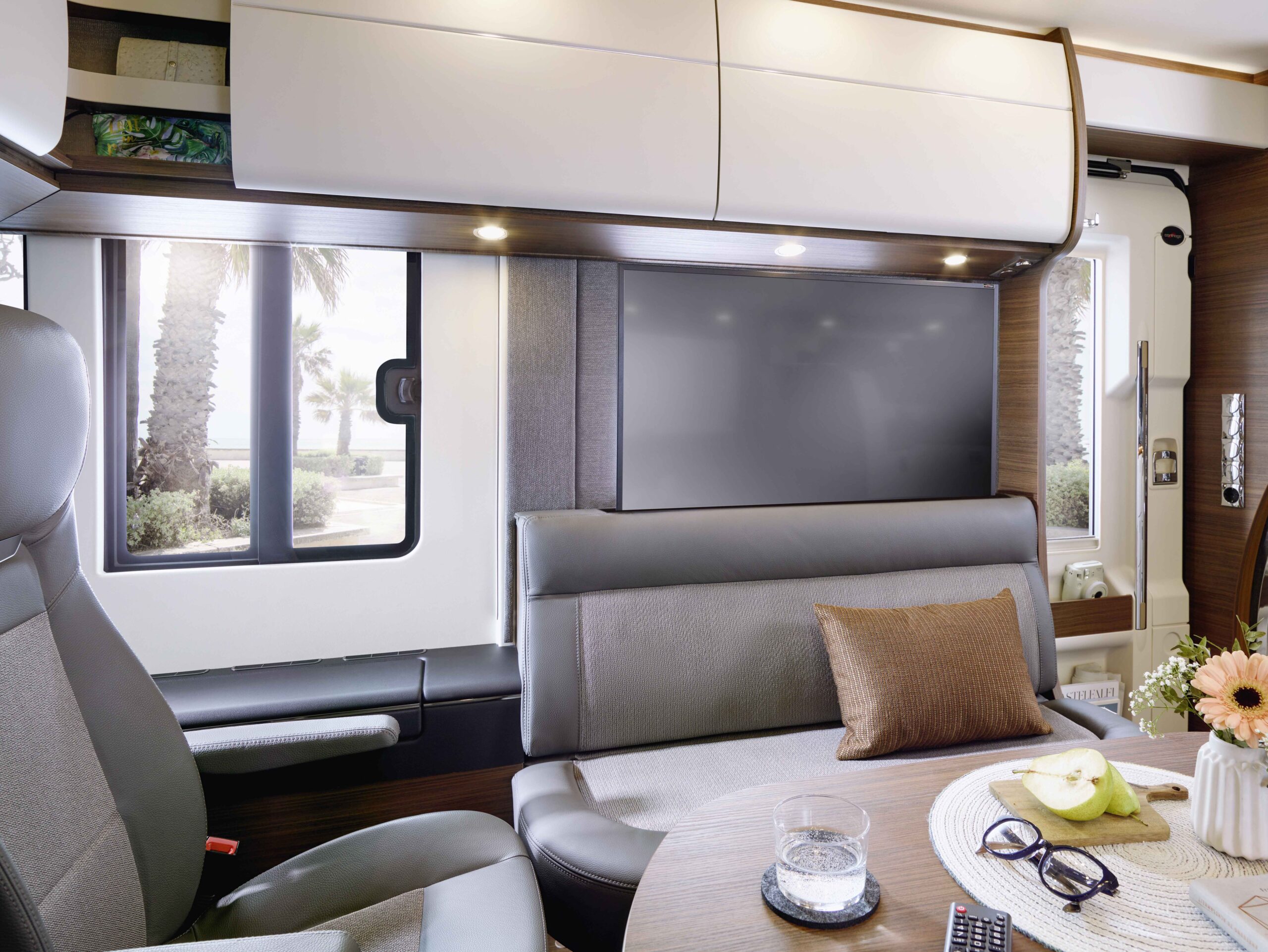 Modernes Wohnmobil-Interieur mit einem Essbereich mit einer gepolsterten Bank und einem Tisch mit Blumen, Obst, einem Glas Wasser und einer Fernbedienung, neben großen Fenstern mit Blick auf Palmen und Sonnenlicht draußen.