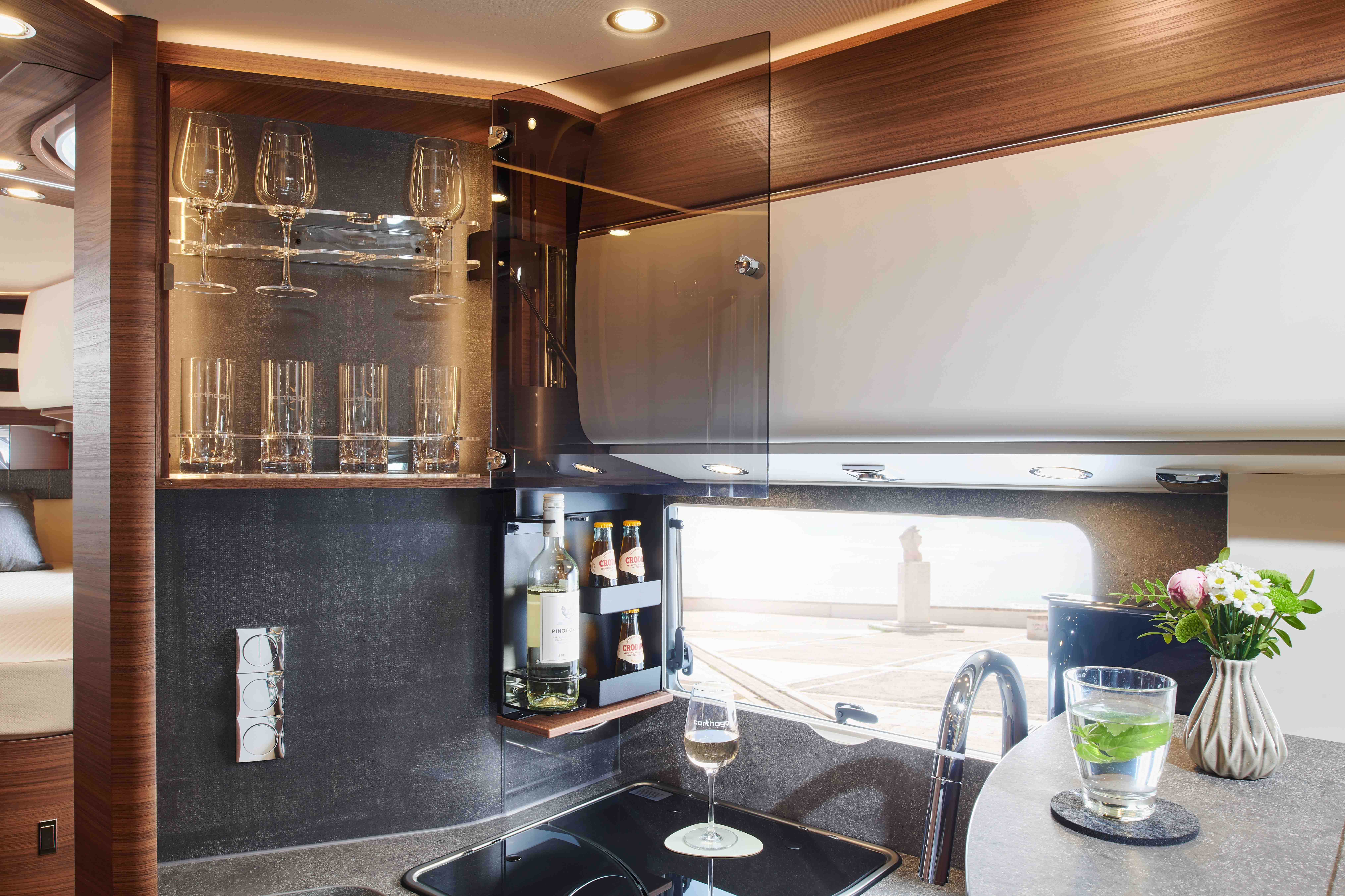 Modern kitchen interior with open glass cabinet displaying wine glasses and tumblers. A wine bottle rack holds bottles beside a window. On the countertop are a wine glass, a glass of water with lime, and a vase with flowers.