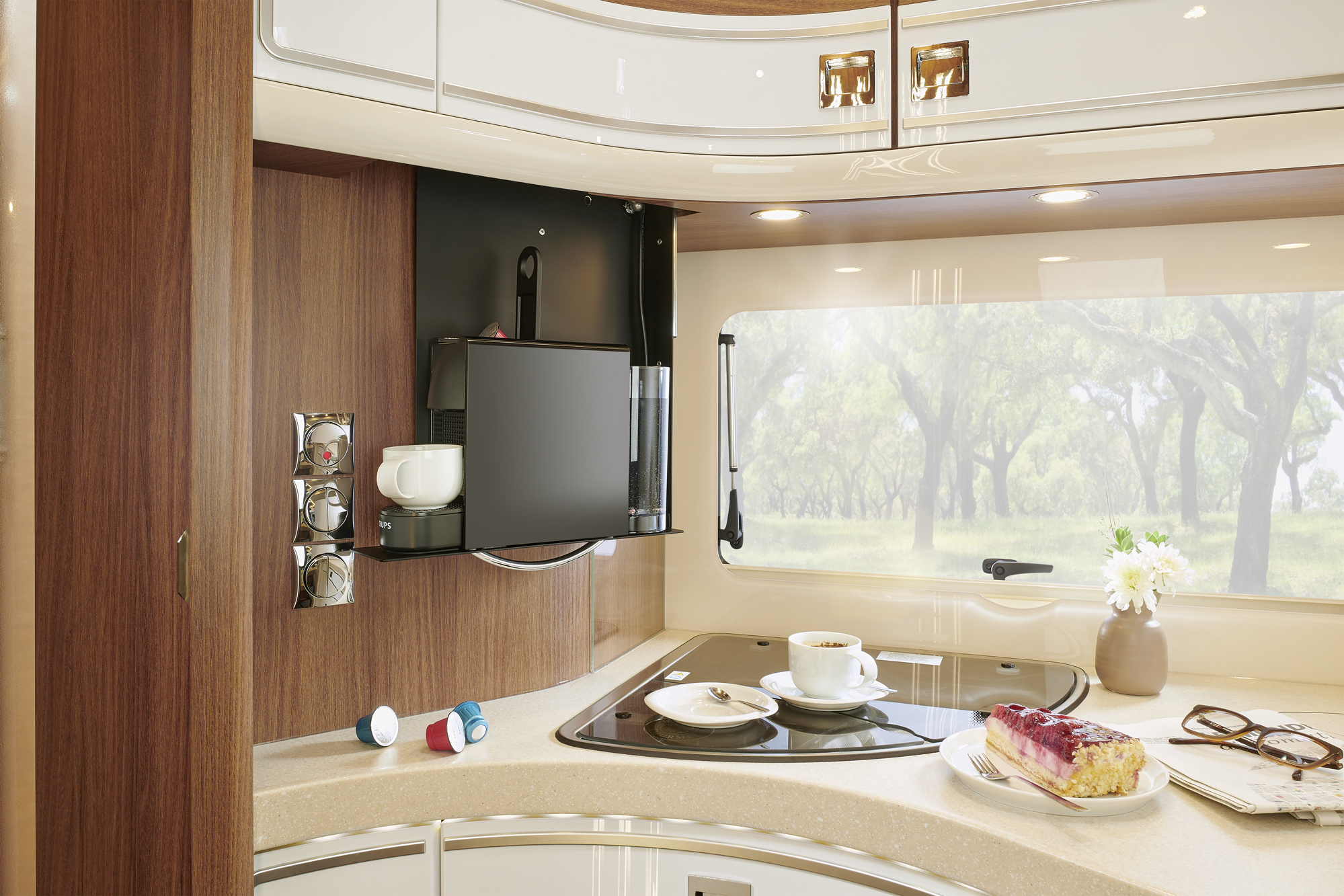 A modern RV kitchen with wooden cabinets, an electric stovetop, a mounted coffee machine with cups, a window showing trees, and a counter with coffee cups, a plate of cake, a vase of white flowers, and coffee pods.