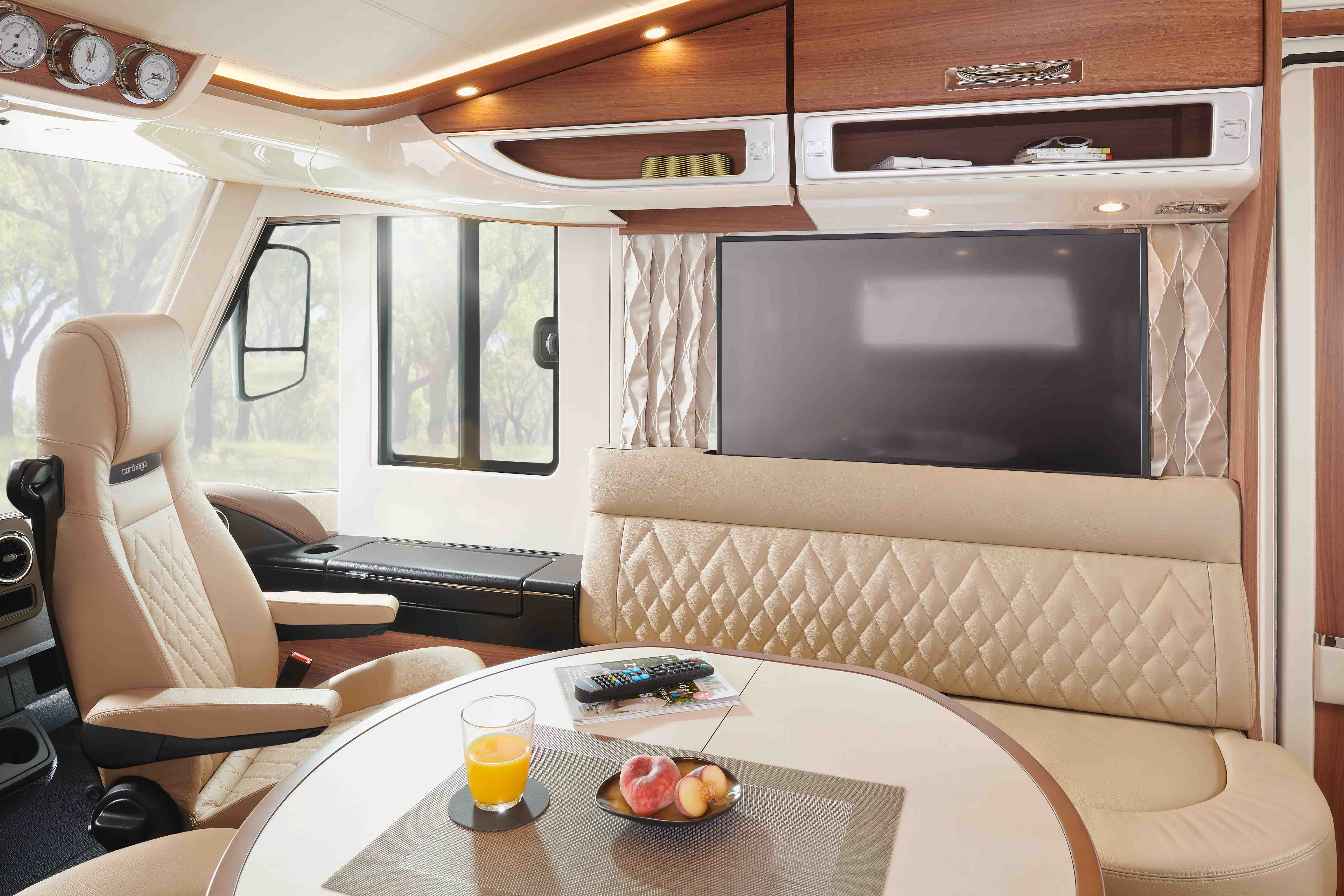 Interior of a modern RV with beige leather seats, a wall-mounted TV, overhead storage, and a dining table set with a remote, orange juice, and fruit. Large windows let in natural light.