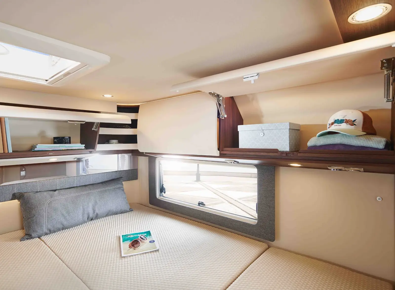 Eine gemütliche Bootskabine mit einem beigen Bett, grauen Kissen, einer Zeitschrift und einem großen Fenster. In den Regalen über dem Bett stehen dekorative Kisten und ein Hut. Mit den Holzakzenten und der sanften Beleuchtung erinnert das Ambiente an den Komfort einer Carthago Chic C Line Einrichtung.