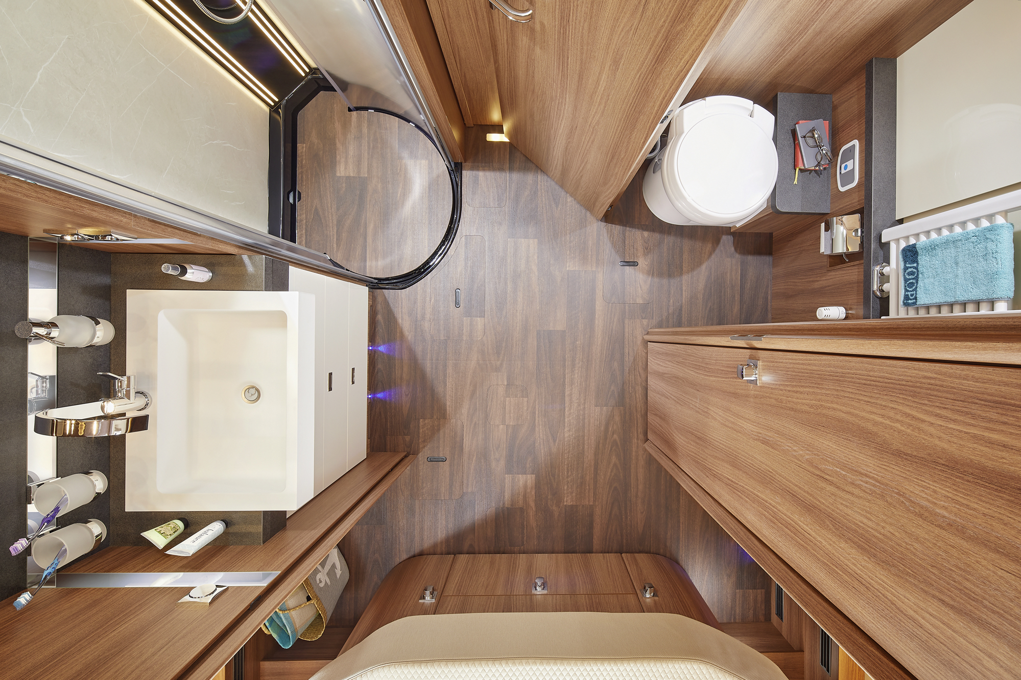 Overhead view of a modern RV bathroom with wood paneling, a white sink, a rounded shower with glass door, a toilet, and organized toiletries on shelves and counters. Towels and accessories add touches of color.