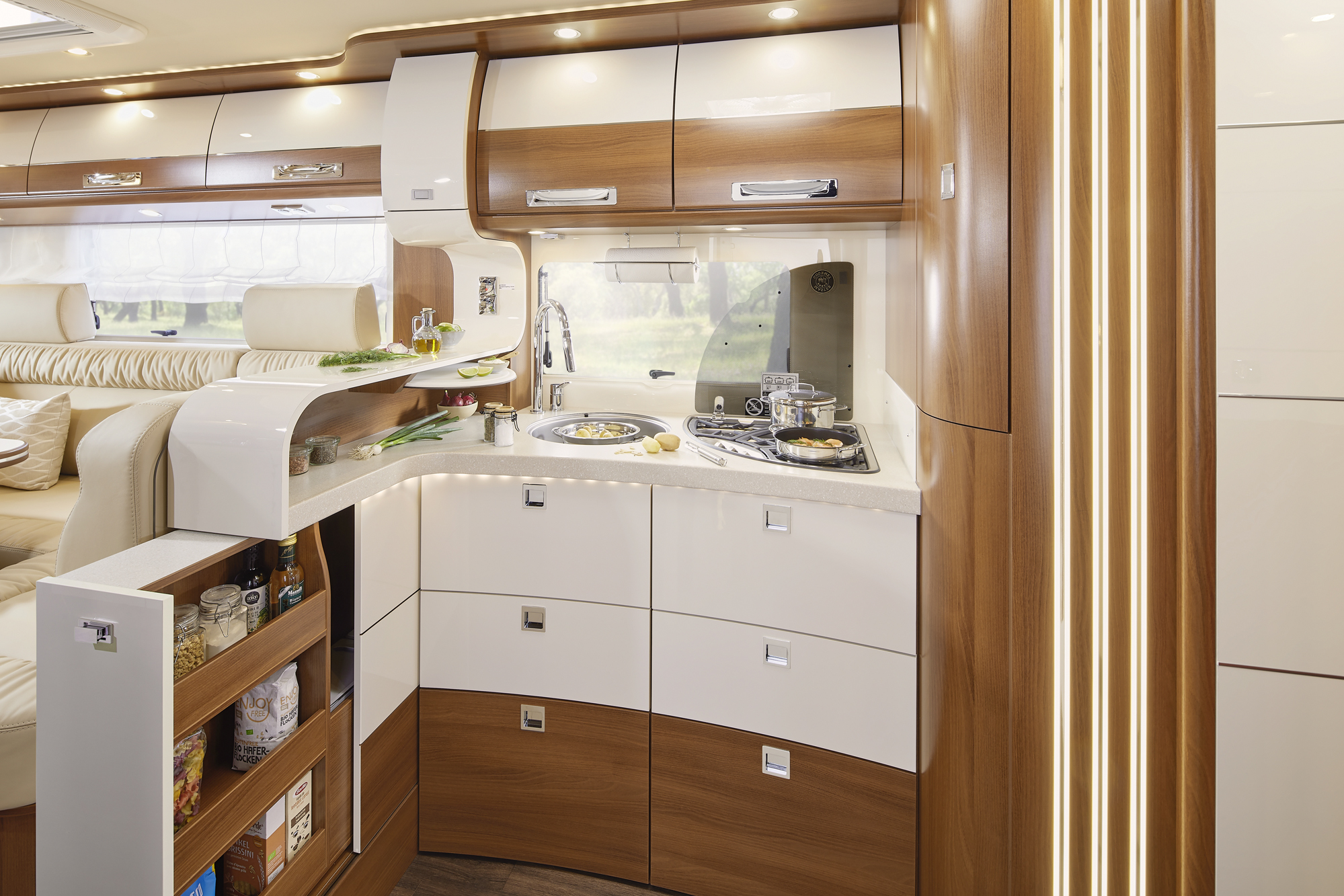 Moderne Wohnmobilküche mit glänzend weißen und hölzernen Schränken, einer kleinen Spüle, einem Herd mit Pfannen und offenen, mit Lebensmitteln gefüllten Vorratsregalen. Helle Beleuchtung und große Fenster schaffen einen gemütlichen und funktionalen Raum.