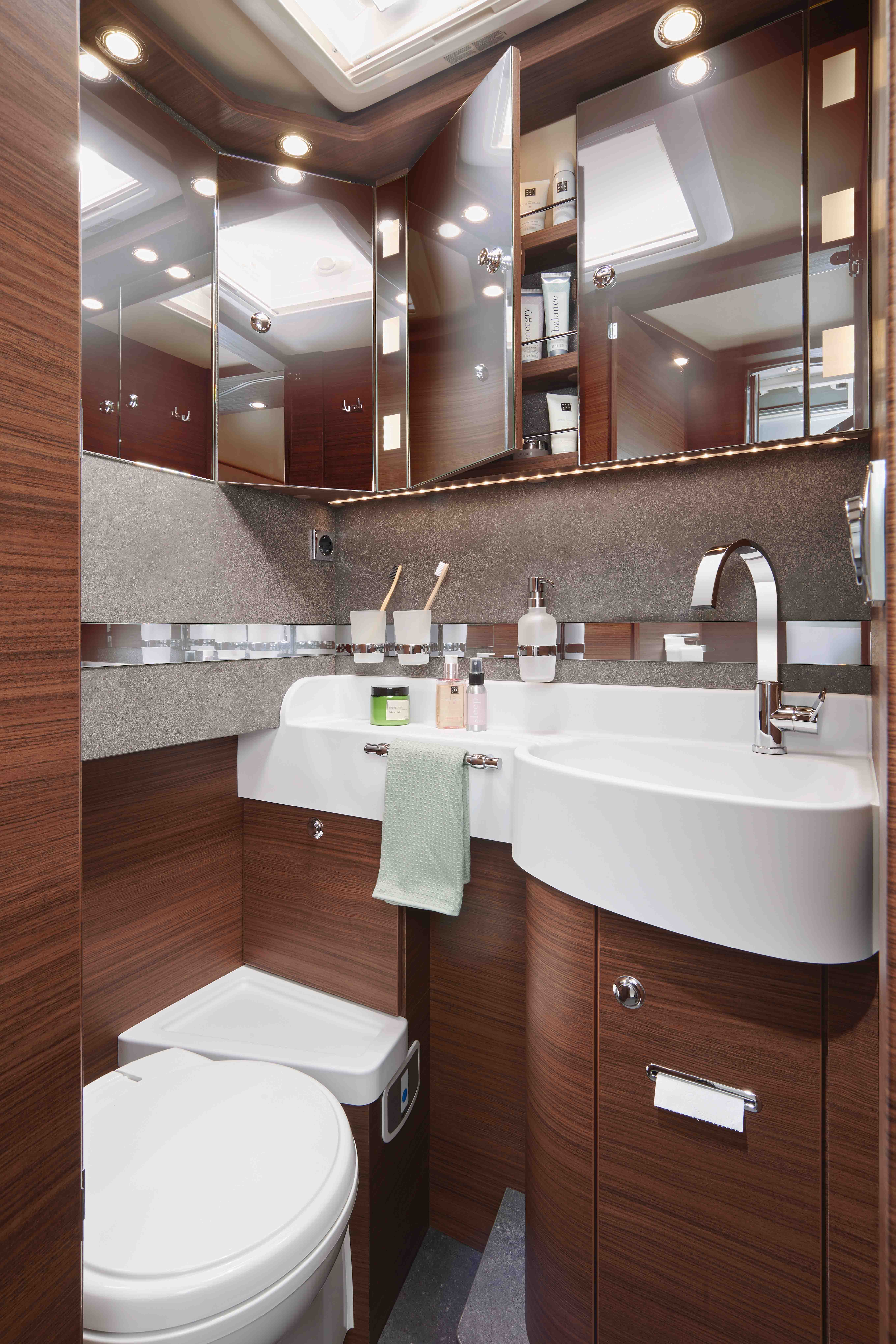 Modern bathroom with wood-paneled cabinets, a white sink with a towel draped over it, a toilet, mirrored storage, and shelves holding toiletries under bright ceiling lights.