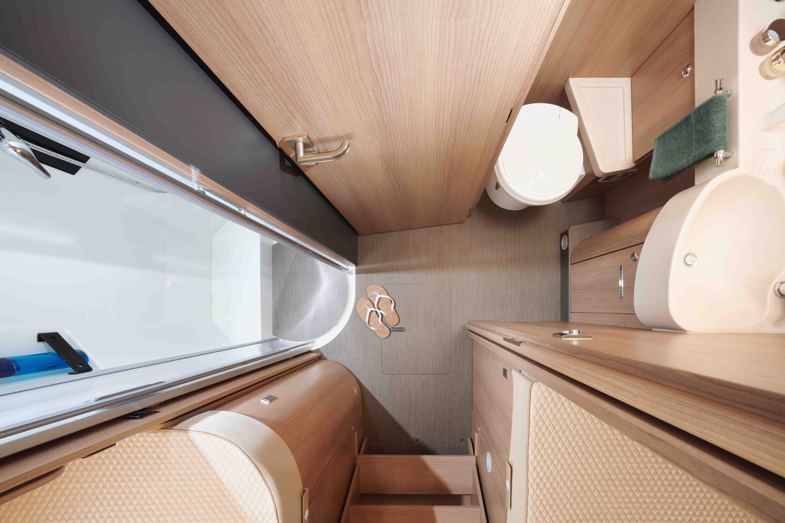 Draufsicht auf ein kompaktes, modernes Wohnmobil-Badezimmer mit Holzschränken, einer weißen Toilette, einem kleinen Waschbecken, Ablageflächen, Hausschuhen auf dem Boden und einem Duschbereich mit einer Glastür.