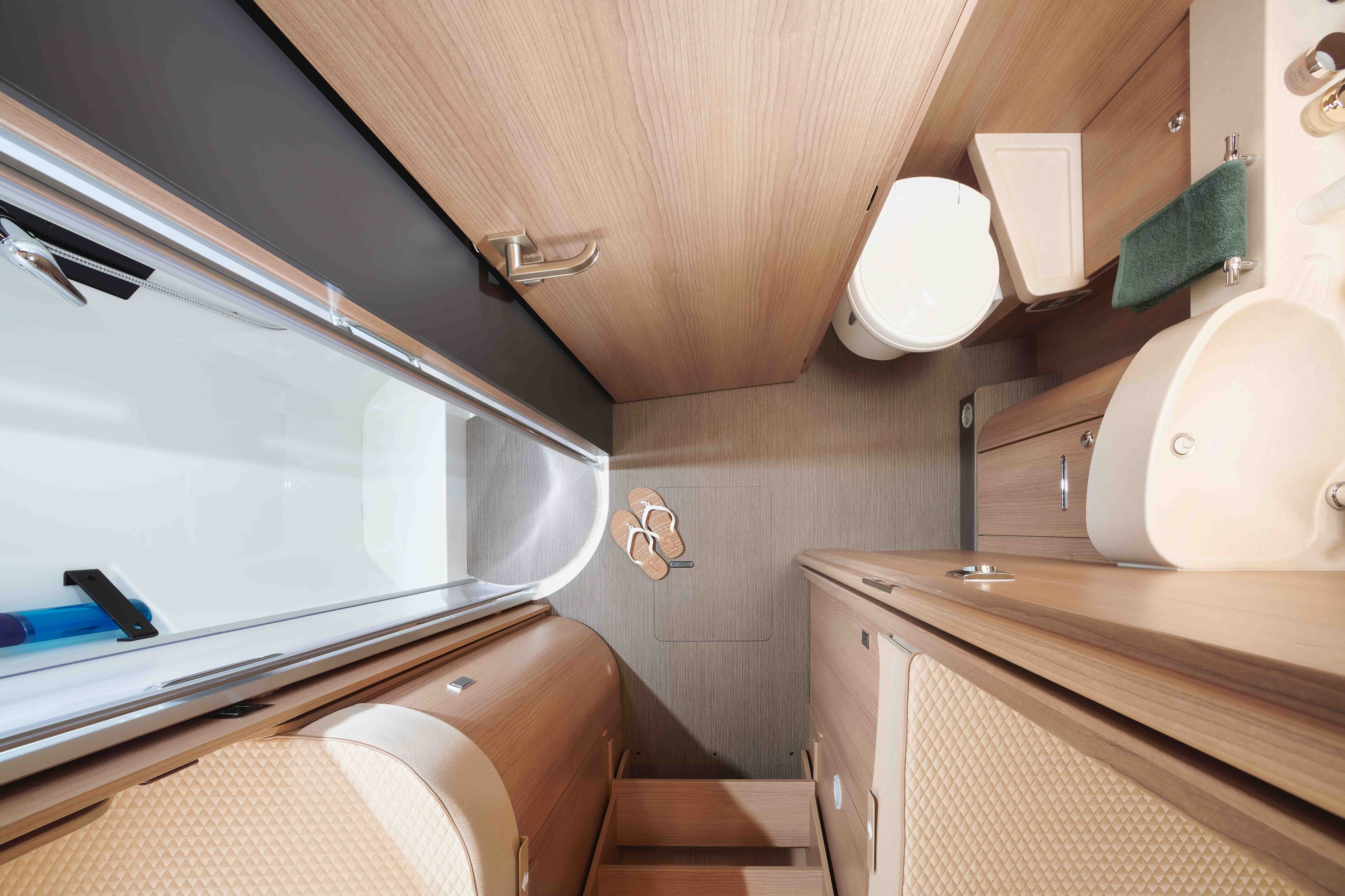 Vue d'ensemble d'une salle de bains compacte aux accents de bois, montrant des toilettes, un lavabo, une douche avec une porte transparente, une serviette et une paire de pantoufles sur le sol. L'espace est bien organisé et moderne.