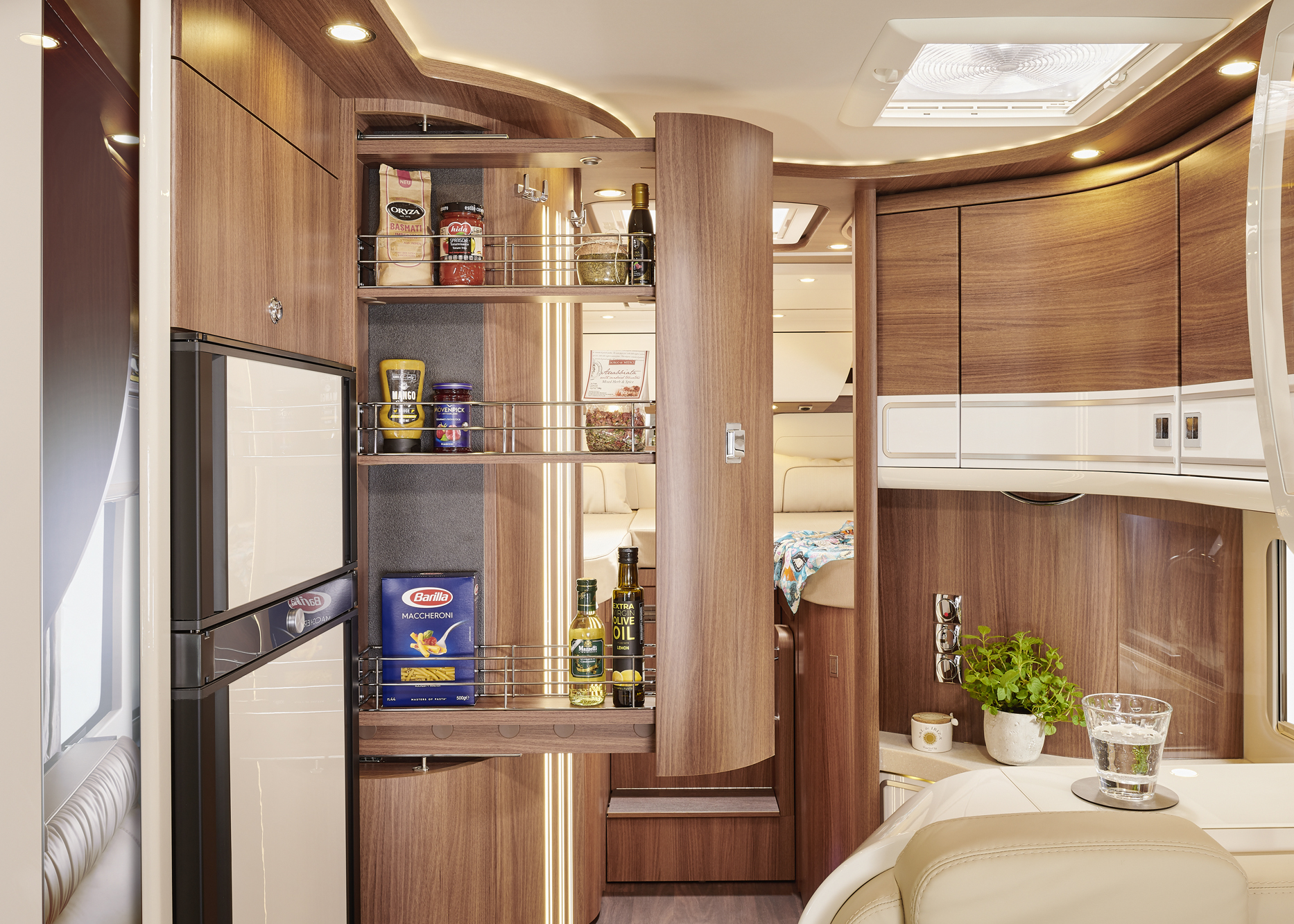 Eine moderne Wohnmobilküche mit Holzschränken, einer offenen, ausziehbaren Speisekammer mit Vorräten, einem Kühlschrank und einem Tresen mit einem Topf mit Kräutern und einem Glas Wasser. Die warme Beleuchtung schafft eine gemütliche Atmosphäre.
