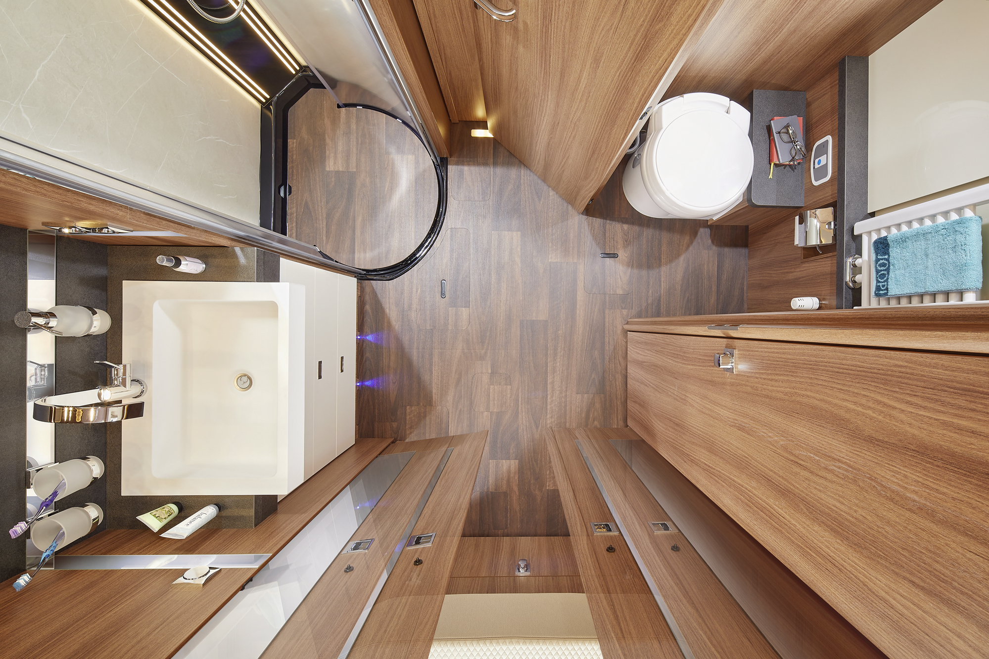 Bovenaanzicht van een moderne badkamer met muren en vloer met houtpatroon, een witte wastafel, douche met glazen deur, toilet, handdoekenrek met blauwe handdoek en toiletartikelen netjes gerangschikt op planken en aanrecht.