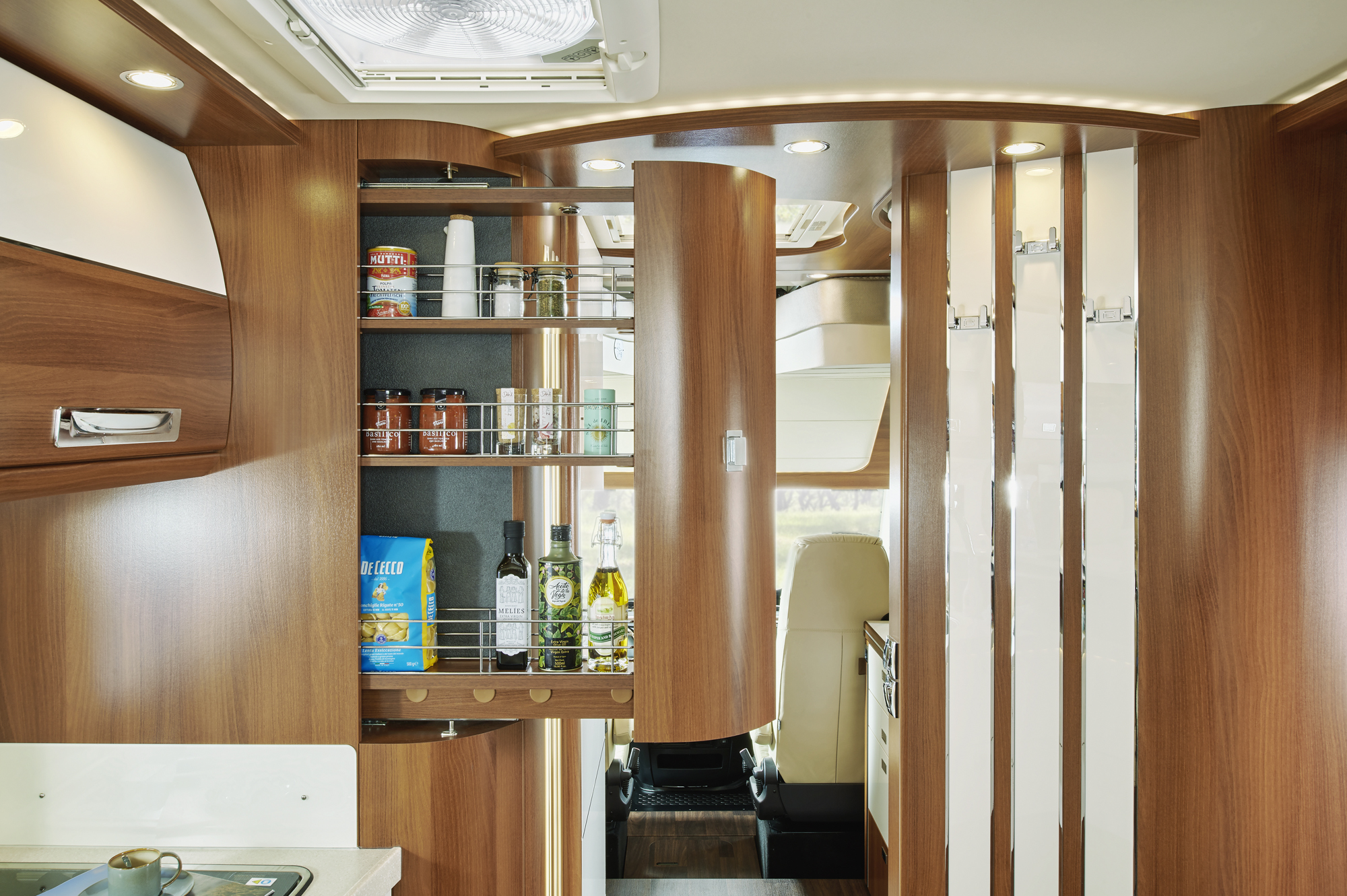 Eine moderne Wohnmobilküche mit Holzschränken und einer ausziehbaren Speisekammer, die mit Lebensmitteln, Flaschen und Dosen gefüllt ist. Der Innenraum ist mit Oberlichtern gut beleuchtet und hat ein schlankes, organisiertes Design.