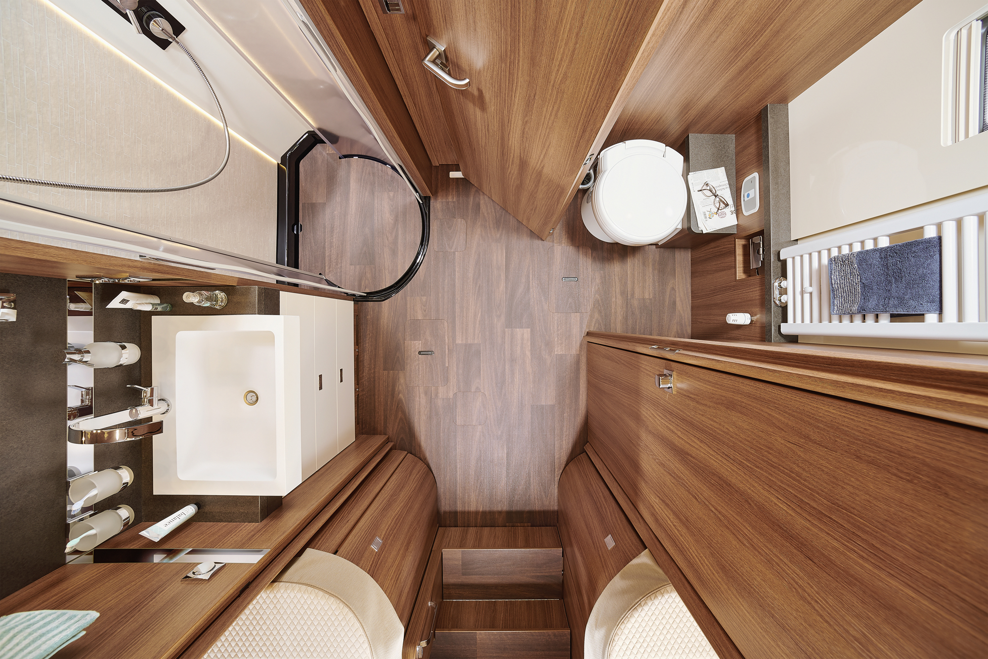 Blick von oben auf ein kompaktes, modernes Wohnmobil-Badezimmer mit Holzverkleidung, einem weißen Waschbecken, einer Toilette und einer Dusche. Der Raum ist aufgeräumt und verfügt über schlichte Armaturen und integrierten Stauraum.