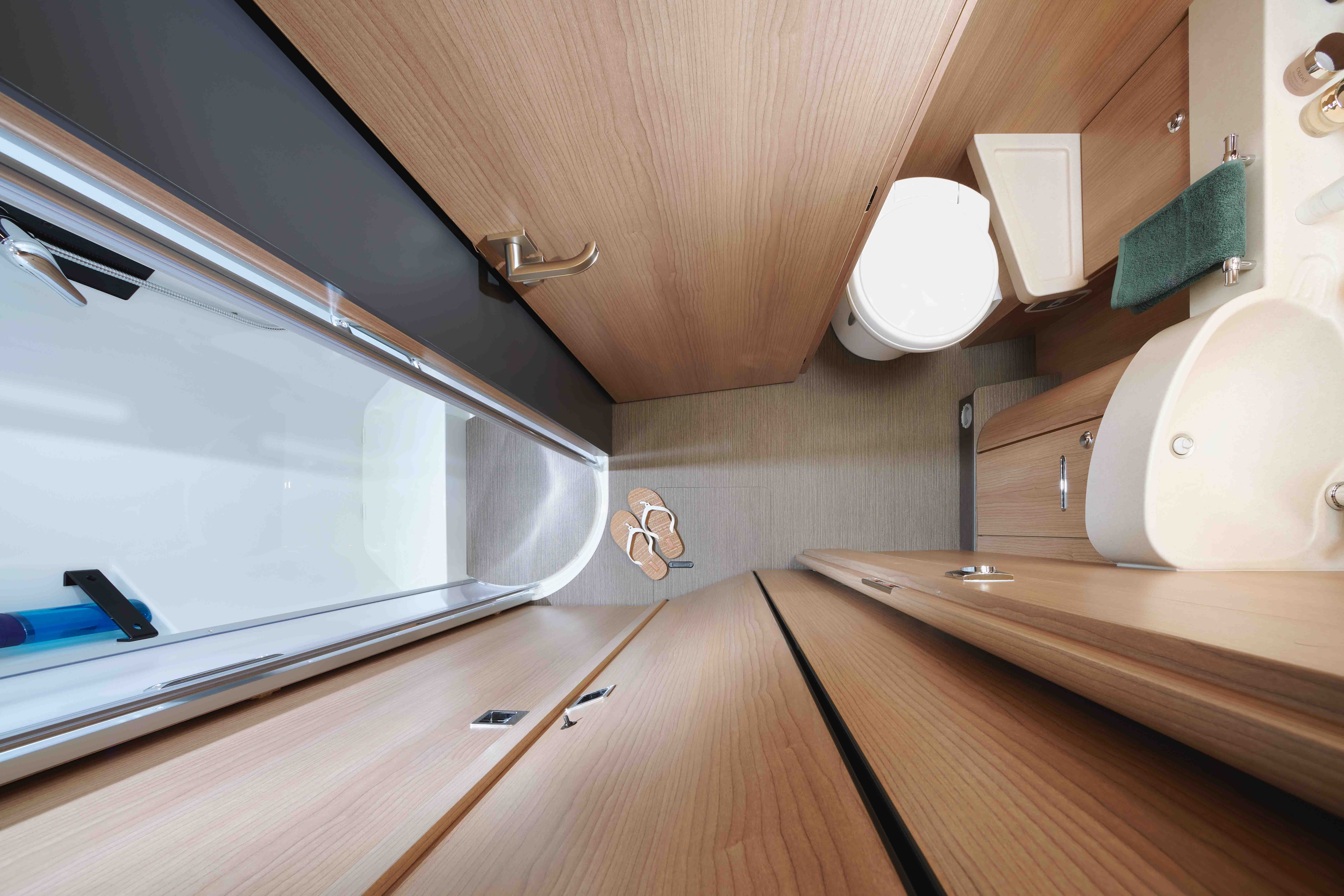 Vue aérienne d'une salle de bain compacte avec des armoires en bois, des toilettes, un petit lavabo, une douche avec une porte en verre, une serviette verte et une paire de pantoufles sur le sol. L'espace est bien organisé.
